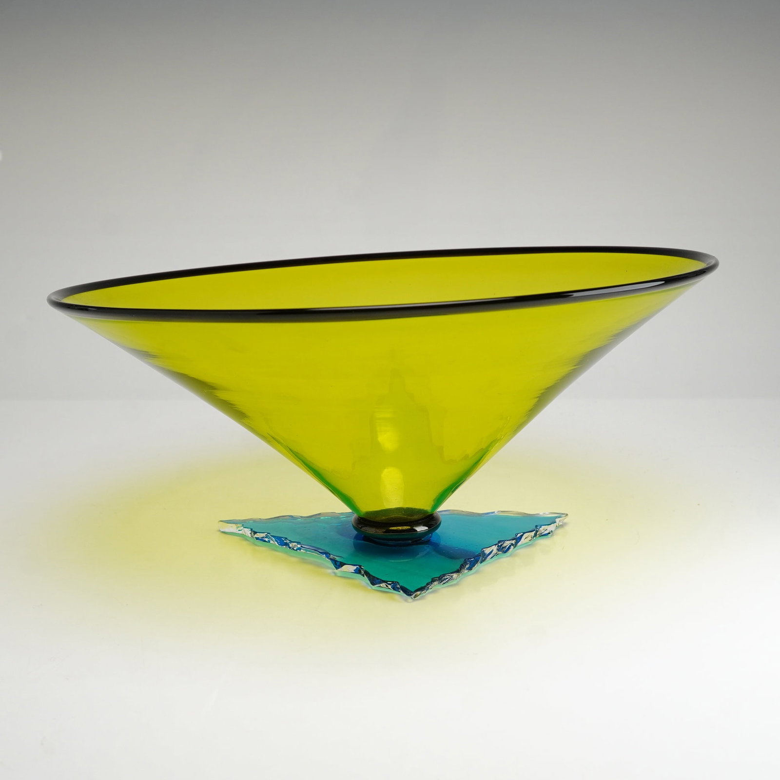Studio Art Glass Bowl with Jagged Edge Base: The vessel features a large conical form with a wide mouth and an asymmetrical slanted rim. The bowl is composed of translucent greenish yellow glass and is finished with a deep amethyst or black lip 