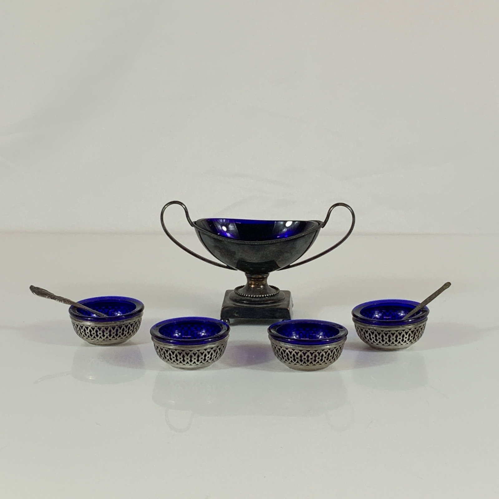 7-Piece Grouping of Sterling Silver and Cobalt Glass Salt Cellars including Gorham and MEKA Denmark: This 7-piece salt cellar grouping consists of an assembled set of sterling silver table articles with cobalt glass inserts. The collection includes one master double-handled salt cellar, four