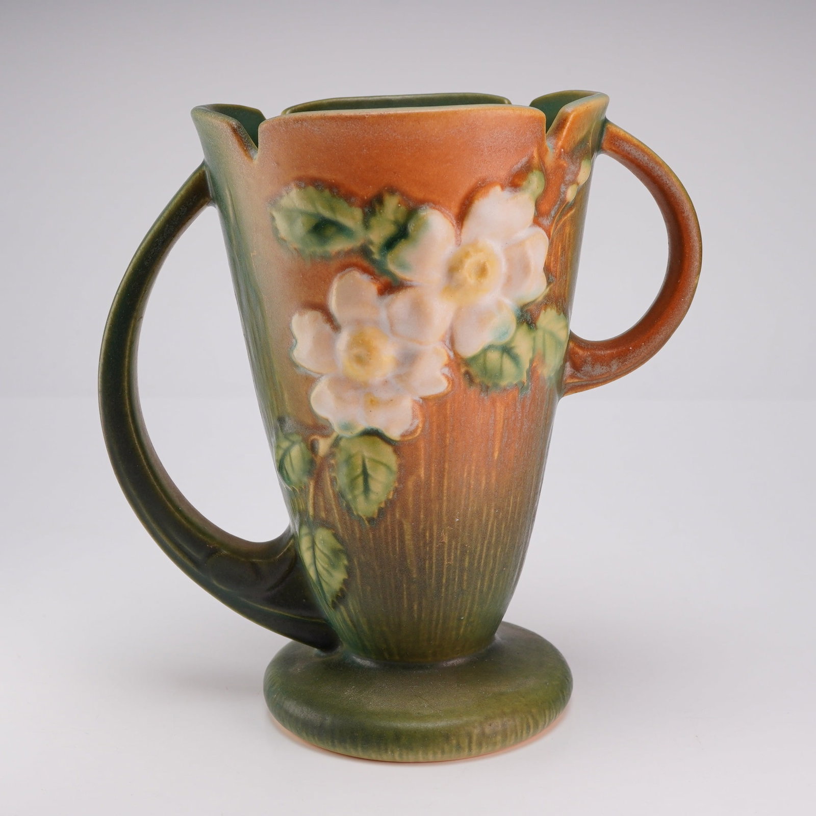 Roseville Pottery Glazed Earthenware Two-Handled Vase, White Rose Brown 982-7: This Roseville Pottery earthenware vase is from the White Rose pattern, introduced circa 1940. The vessel features a footed form with an asymmetrical scalloped rim and two large arching handles that