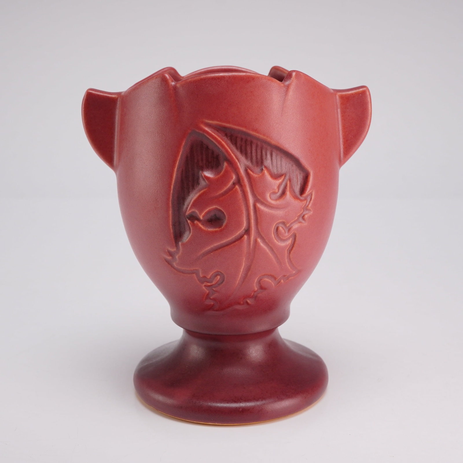 Roseville Silhouette Russet Red Art Pottery Vase: This is a footed earthenware vase produced by Roseville Pottery in the Silhouette pattern, introduced circa 1950. The piece features a matte russet red glaze over a body decorated with a recessed,
