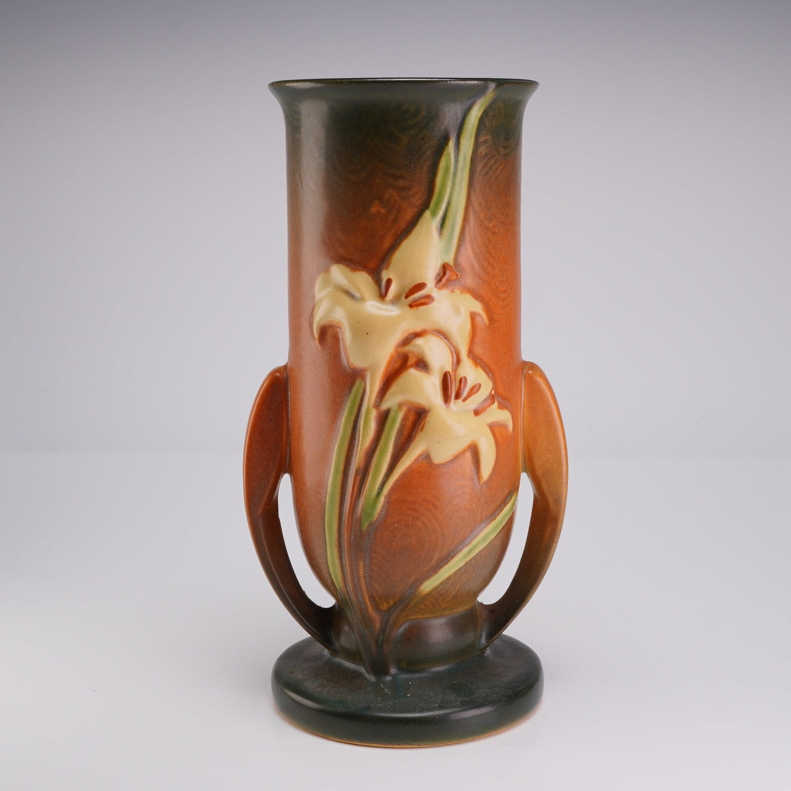 Roseville Pottery Vase Zephyr Lily Brown 145-8: This tall, cylindrical vessel features a slightly flared rim and a circular, tiered foot. The form is distinguished by two wing-like handles that curve upward from the base to the mid-section of the b