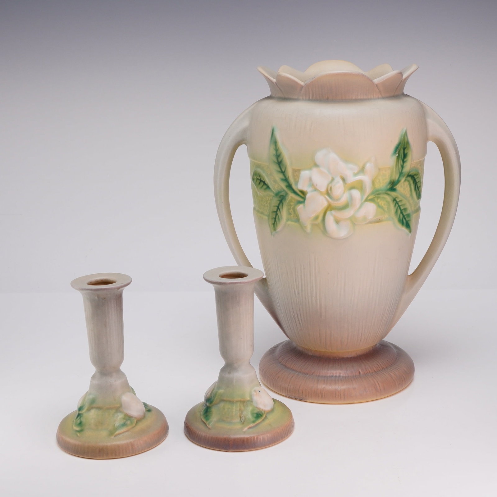 Set of Three Roseville Pottery Gardenia Gray Vase and Candleholders: This three-piece set of American art pottery from Roseville Pottery of Zanesville, Ohio, features the Gardenia pattern, which was introduced in 1950. The set consists of a large twin-handled vase and 