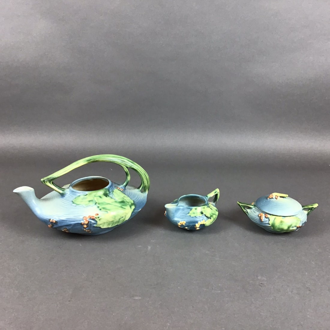Three Piece Roseville Pottery Glazed Tea Service, Bittersweet Blue: A trio of Roseville Pottery earthenware vessels in the Bittersweet pattern, produced circa 1940. The set consists of a teapot, a creamer, and a lidded sugar bowl. Each piece features relief-molded dec