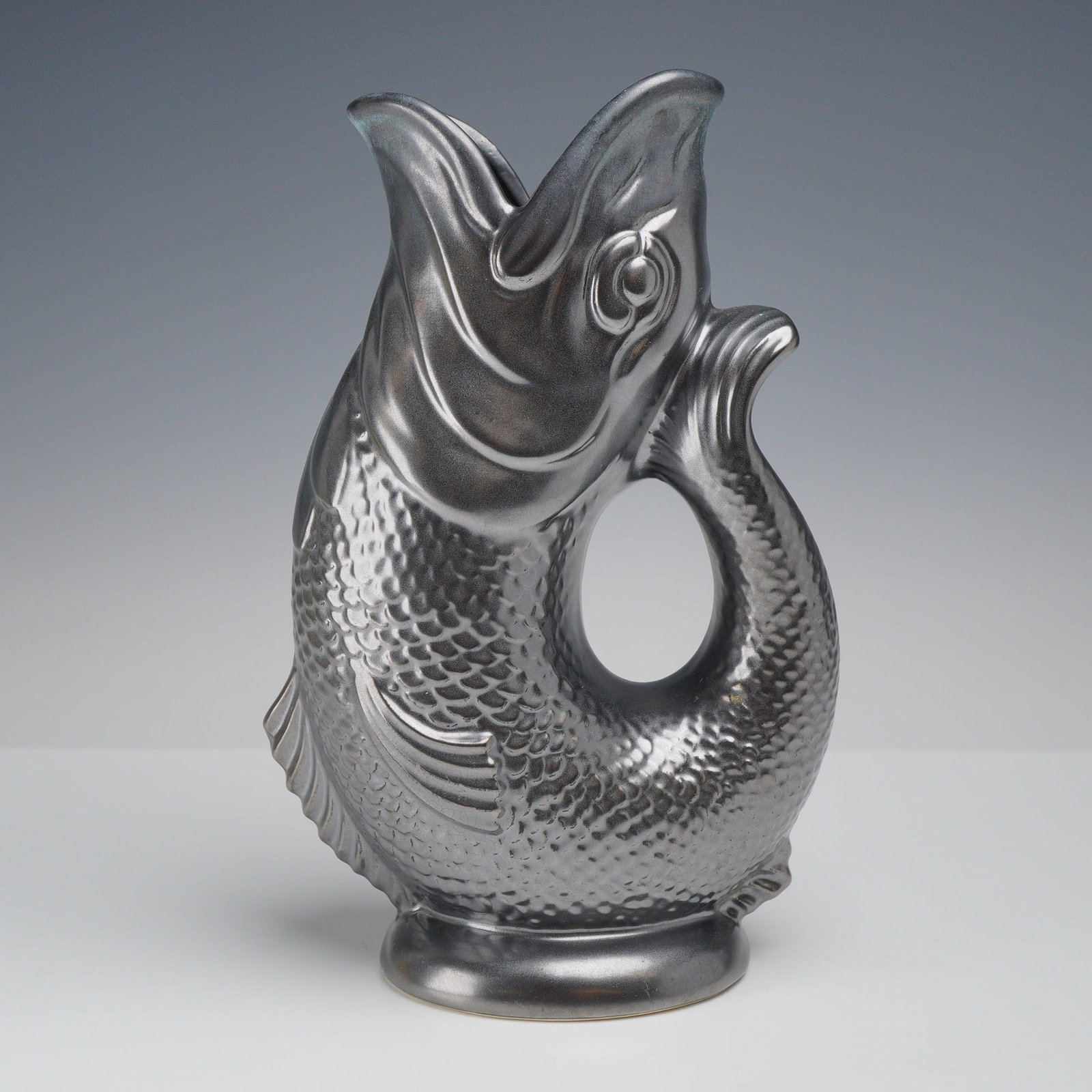 Dartmouth Pottery Metallic Luster Glaze Gurgle Fish Jug: This ceramic pitcher, a part of the Rubin Collection, is a gurgle jug produced by Dartmouth Pottery in Devon, England. The vessel is modeled in the form of a fish, where the open mouth serves as the s