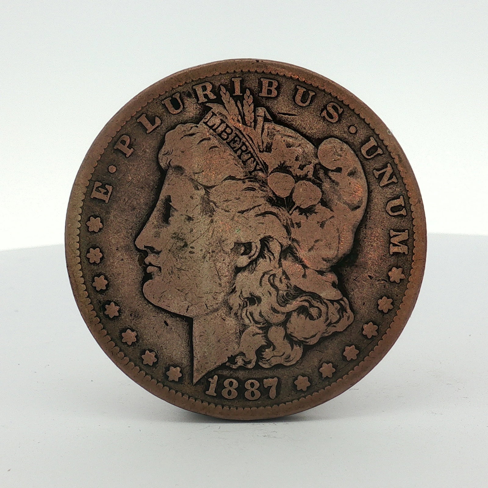 1887-S US One Dollar Morgan Silver Dollar, Circulated: This 1887-S Morgan silver dollar design was minted in San Francisco. The obverse features the date 1887 and the reverse displays an S mint mark for the San Francisco mint below the wreath. The surface