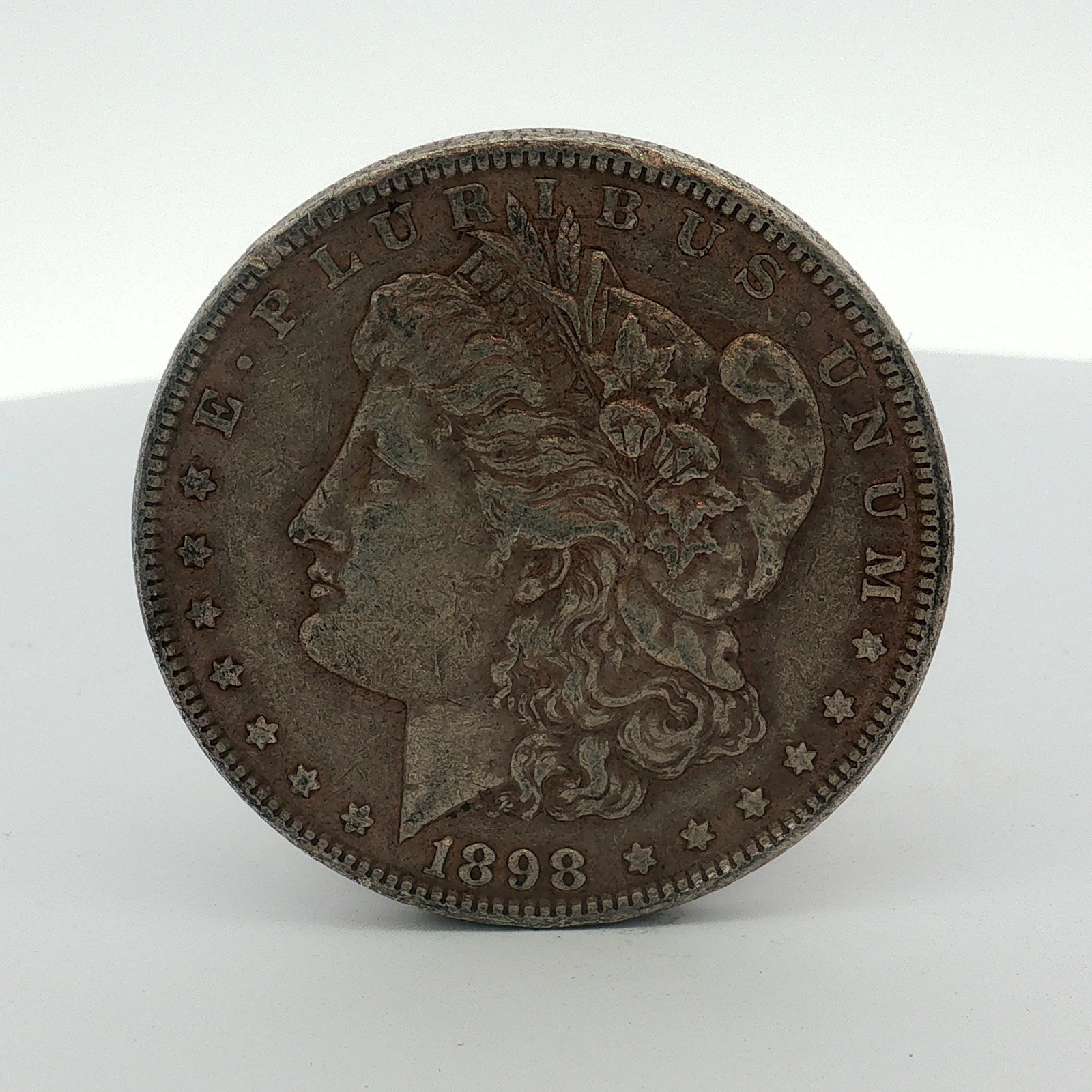 1898 US Morgan Silver Dollar Coin Circulated: This 1898 Morgan silver dollar is a silver coin struck at the Philadelphia Mint. The obverse features the Liberty Head design type, and the reverse features the Eagle with Wreath design type. Inscript