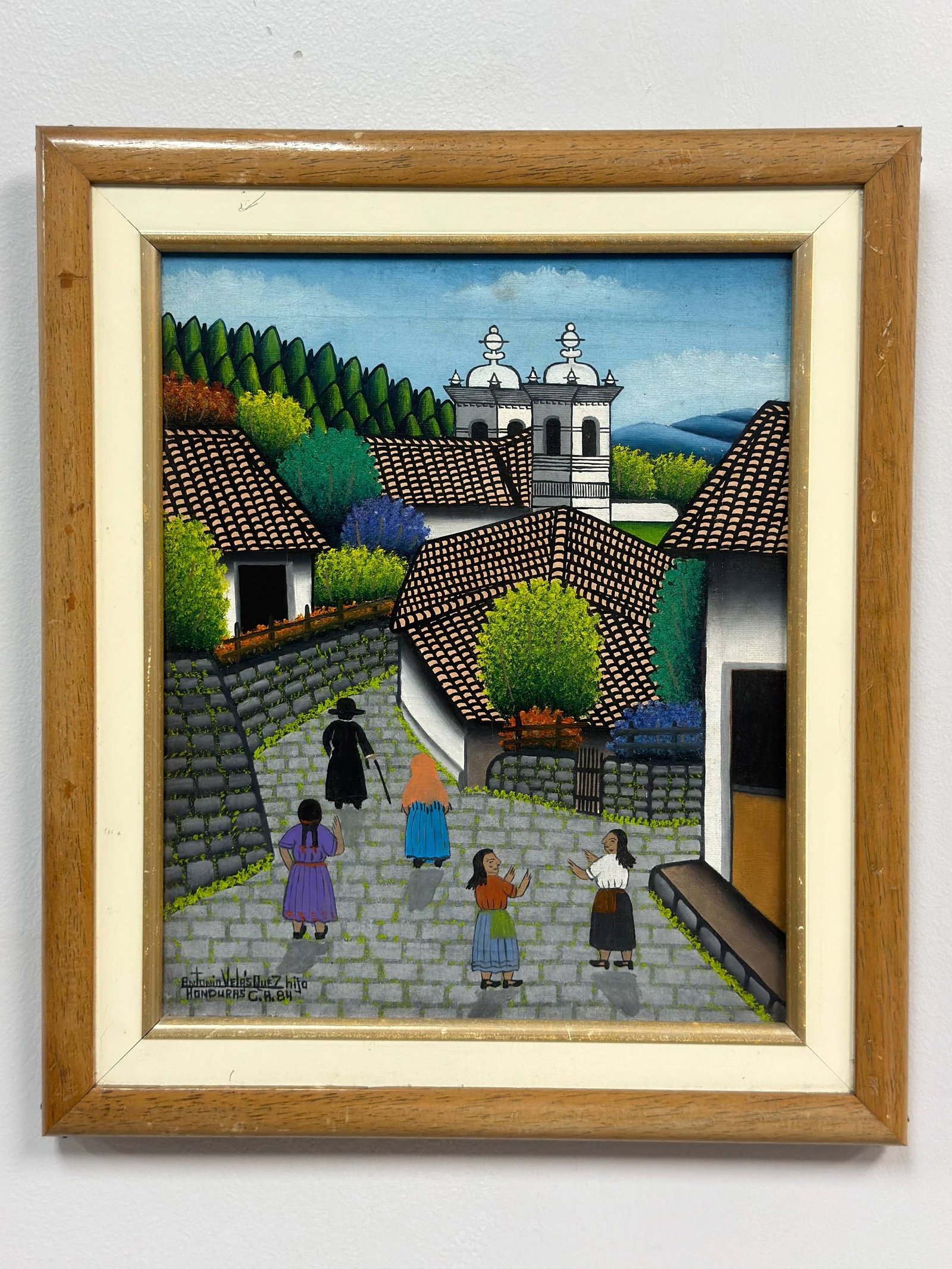 Jose Antonio Velasquez (Honduran, 1906-1983), Village Street Scene: This framed original street scene painting on canvas depicts a traditional village in Honduras, executed in a vibrant naive style. The composition features a central cobblestone path populated by seve