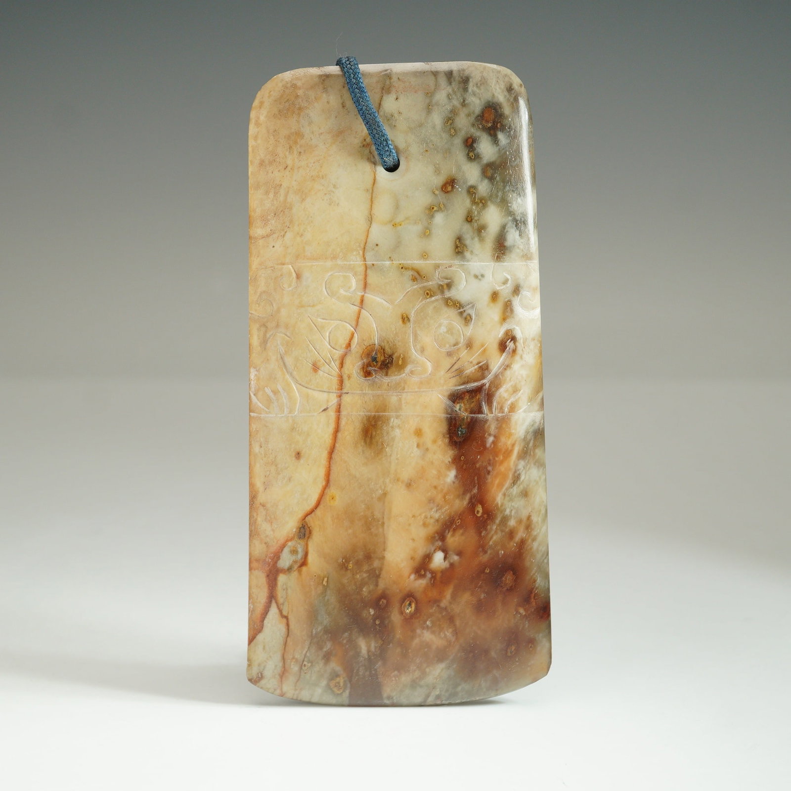 Chinese Archaic-Style Nephrite Jade Blade: Featuring a incised mask design carved in low relief across one side. The blade is fashioned from a mottled stone displaying natural variations in color, including shades of beige, brown, and russet w
