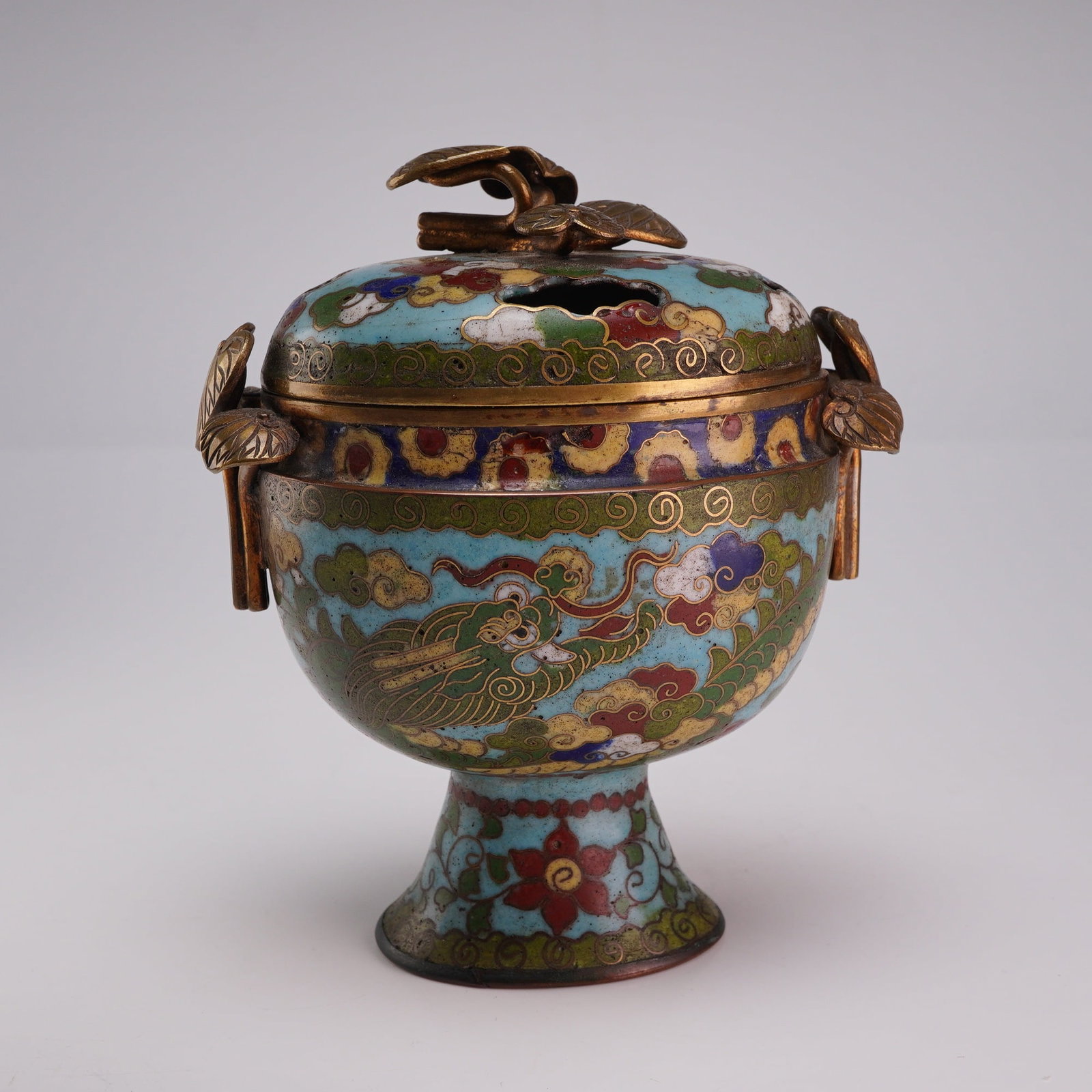 Small Cloisonne Censer and Cover, Dragon and Clouds: A small gold-tone metal footed vessel with a lid featuring cloud-shaped openings, and leaf-shaped handles and finial. The exterior is decorated in cloisonne depicting dragons, clouds, and flowers. Dim
