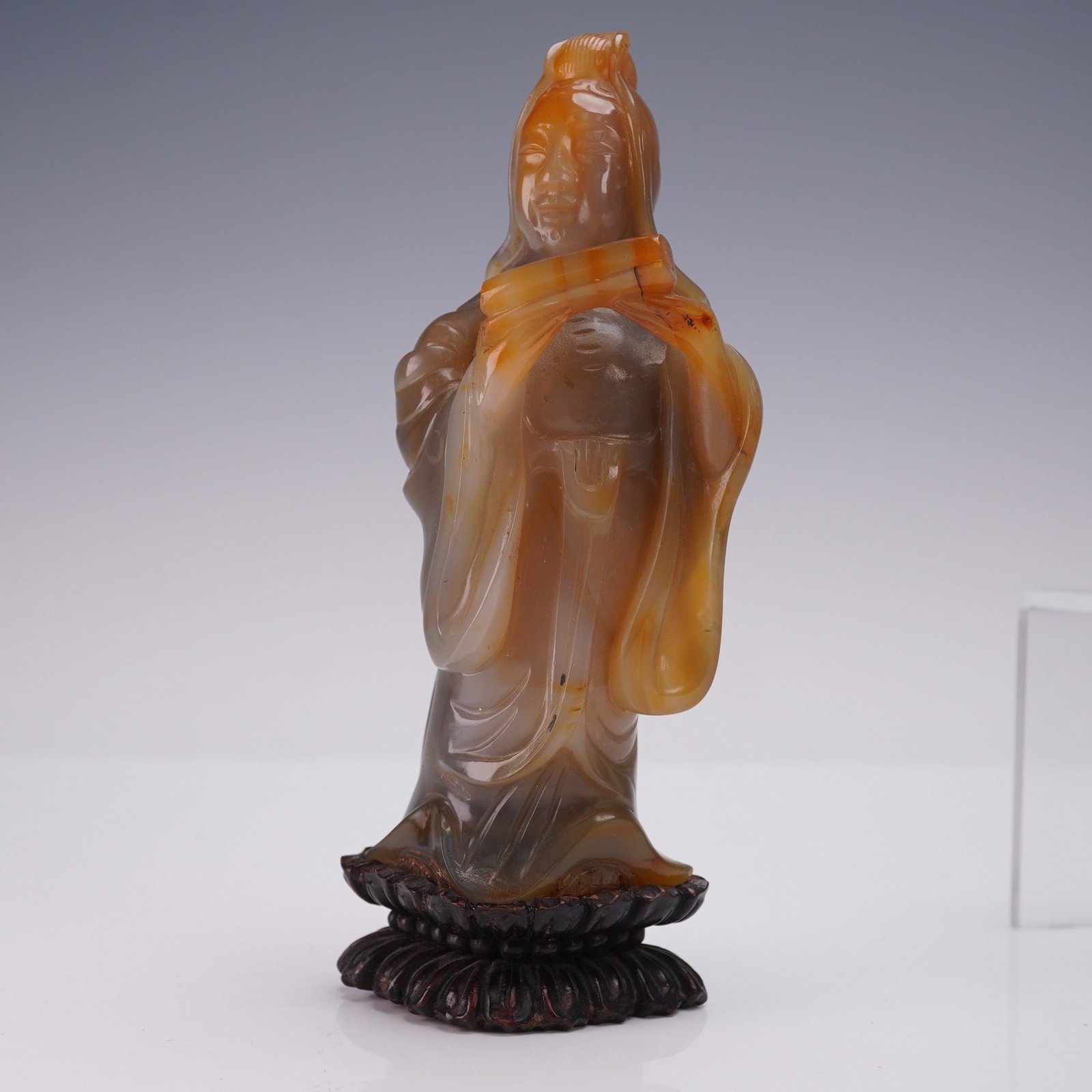 Chinese Agate Guanyin Holding Scroll on Wood Stand: Chinese agate carving of Guanyin holding a scroll, presented on a carved wooden stand. The figure is fashioned from agate displaying natural color variations from light honey to deep amber. Depicted