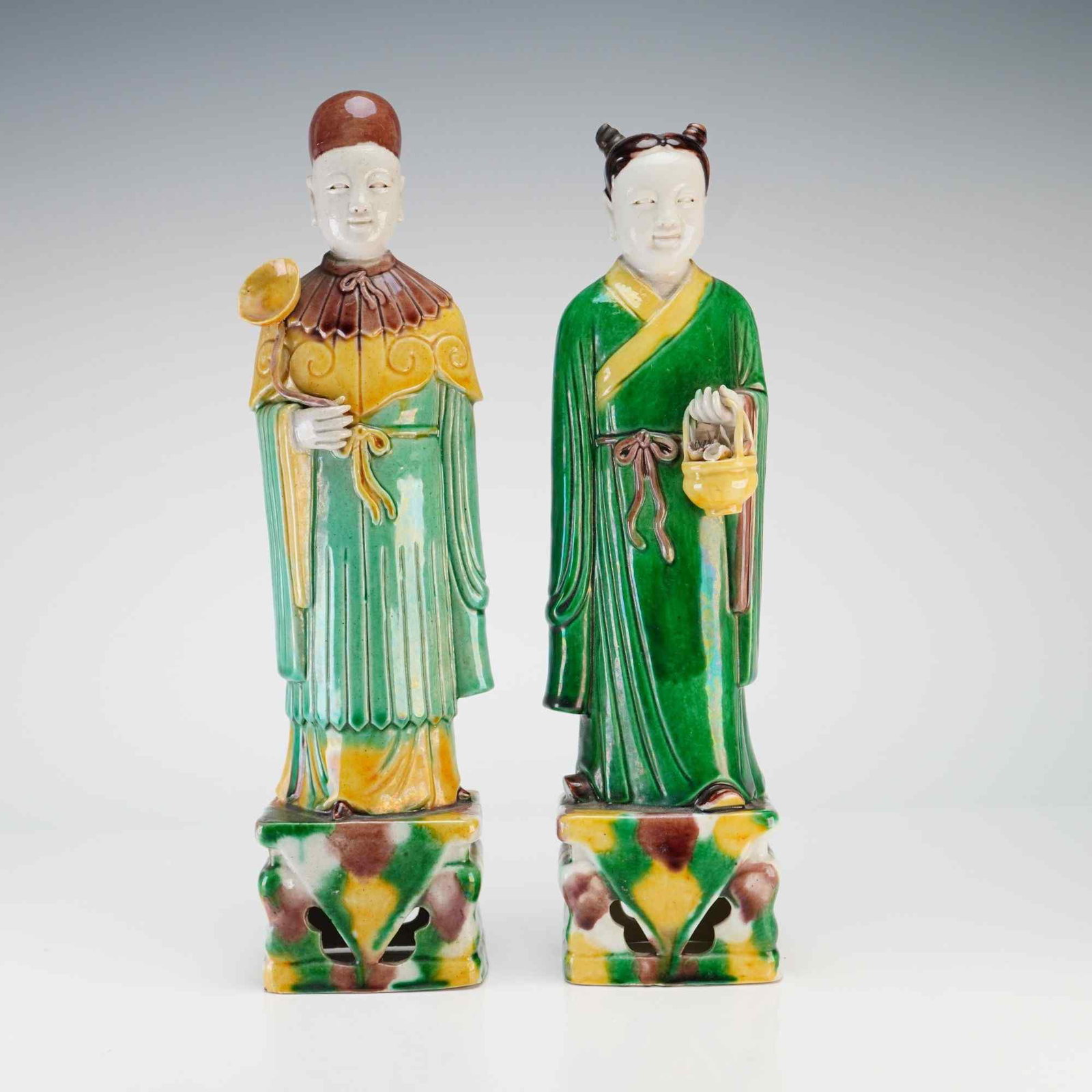2pc Chinese Sancai Glaze Porcelain Figures: Two Chinese porcelain figurines depicting robed individuals standing on a square base, finished in green, yellow, and brown hues. Dimensions Each: 3"L x 2"W x 8.25"H Weight 1.110 lb