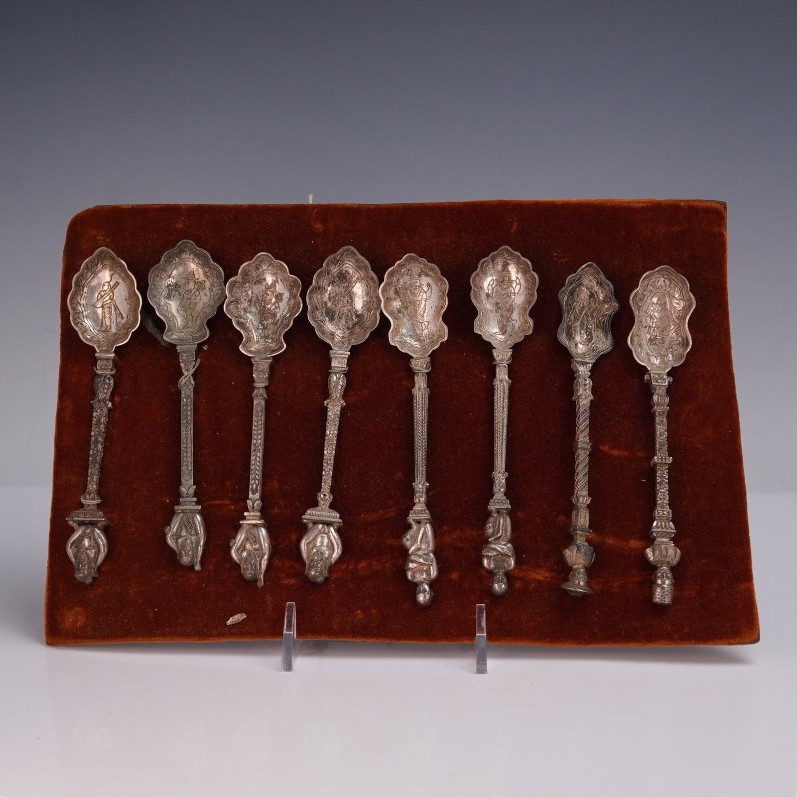 8pc Thai Silver Decorative Spoons: Set of eight Thai silver spoons displayed on a velvet board, each featuring an intricately cast handle topped with a detailed figure and a scalloped bowl engraved with traditional scenes and motifs. T