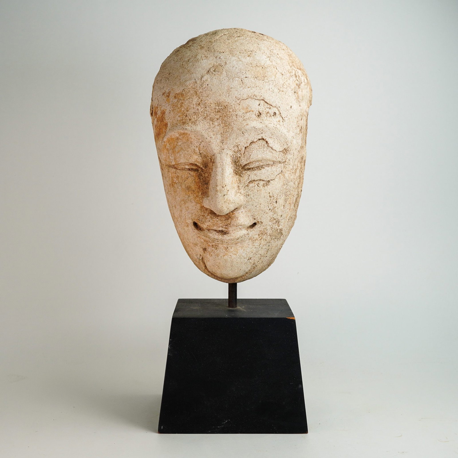 Antique Southeast Asian Carved Stone Funerary Mask: Carved stone face fragment depicting a serene face with closed eyes and a faint smile, mounted on a black display base via a metal rod. The fragment, likely representing a deity or meditative figure,