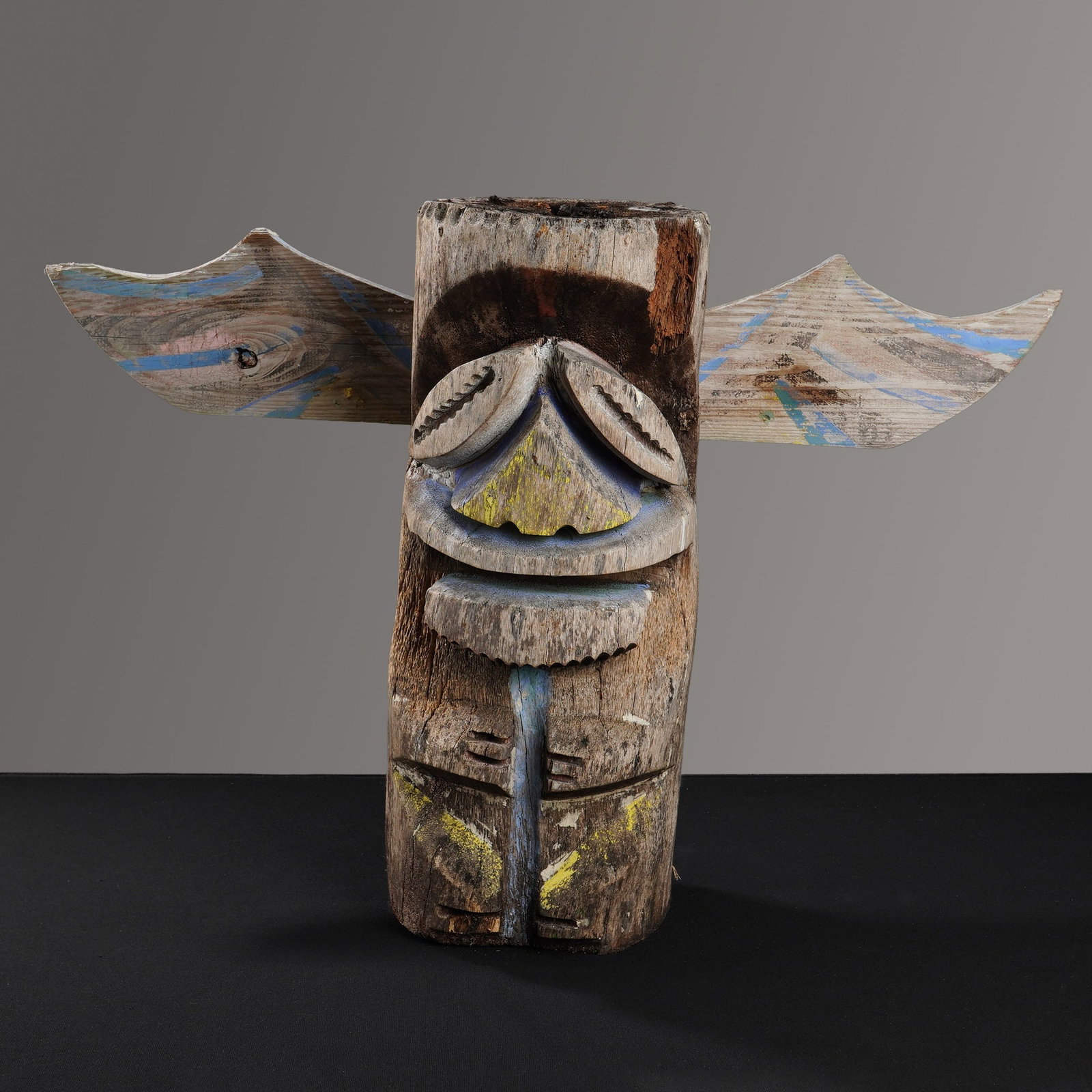 Hand Carved Wooden Winged Totem: This hand carved wooden winged totem is fashioned from a weathered log and features a stylized face with large cowrie-shell-shaped eyes, a triangular nose, and a wide serrated mouth. The figure has a