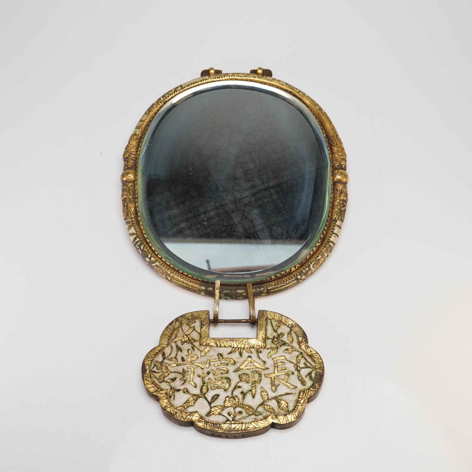 Gilt Copper and Enamel Mounted Mirror with Baby Lock: An oval gilt copper mirror with cream-toned enamel, adorned with a scalloped decorative pendant hanging from the frame. The reverse of the mirror is decorated with branches and leaves, while the