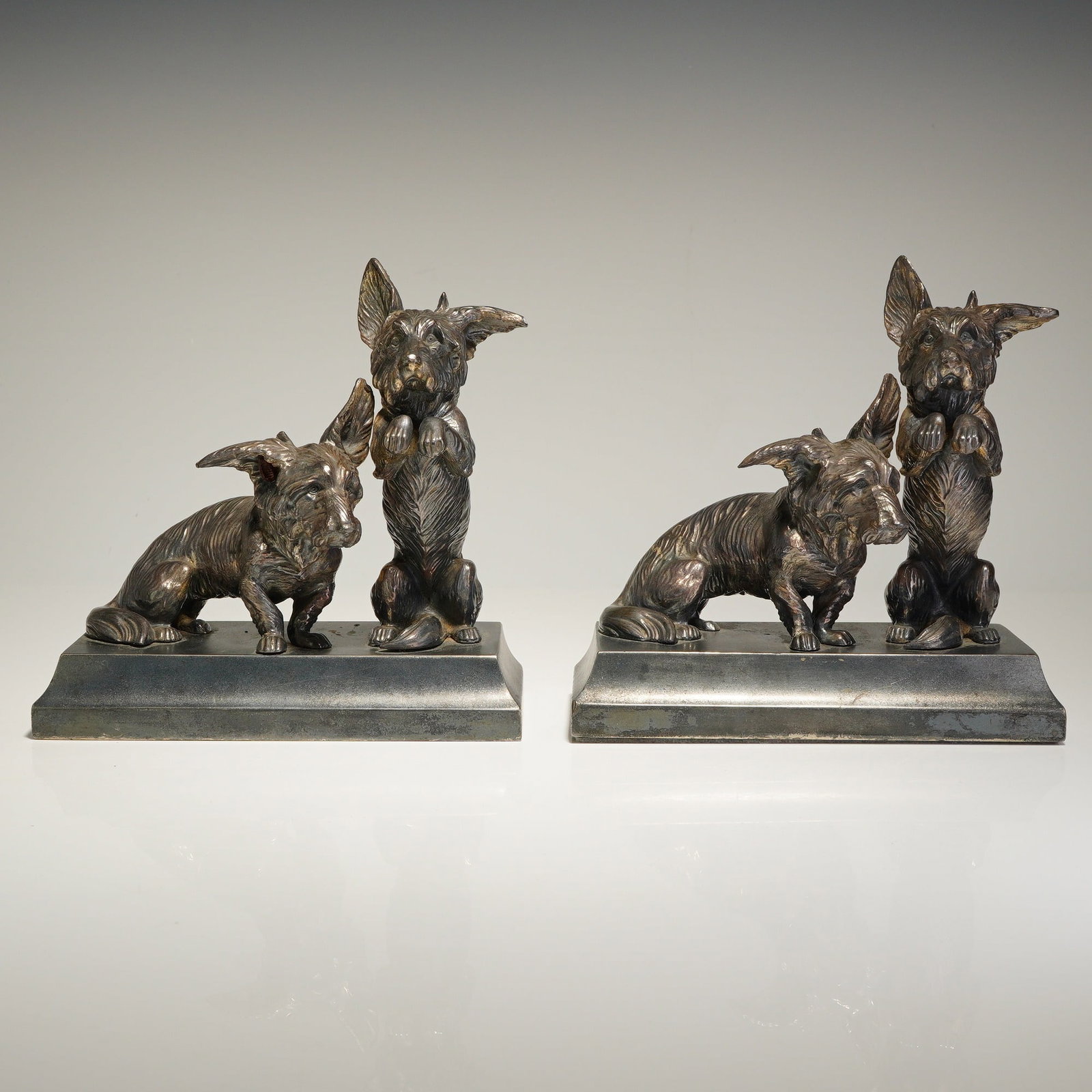 Pair of Jennings Bros Cast Metal Scottish Terrier Dog Bookends: Each item depicts two dogs, one seated and one in a begging posture, mounted on a rectangular beveled base. The figures are finished in a silver-toned metallic coating with a darkened patina. Dimensio