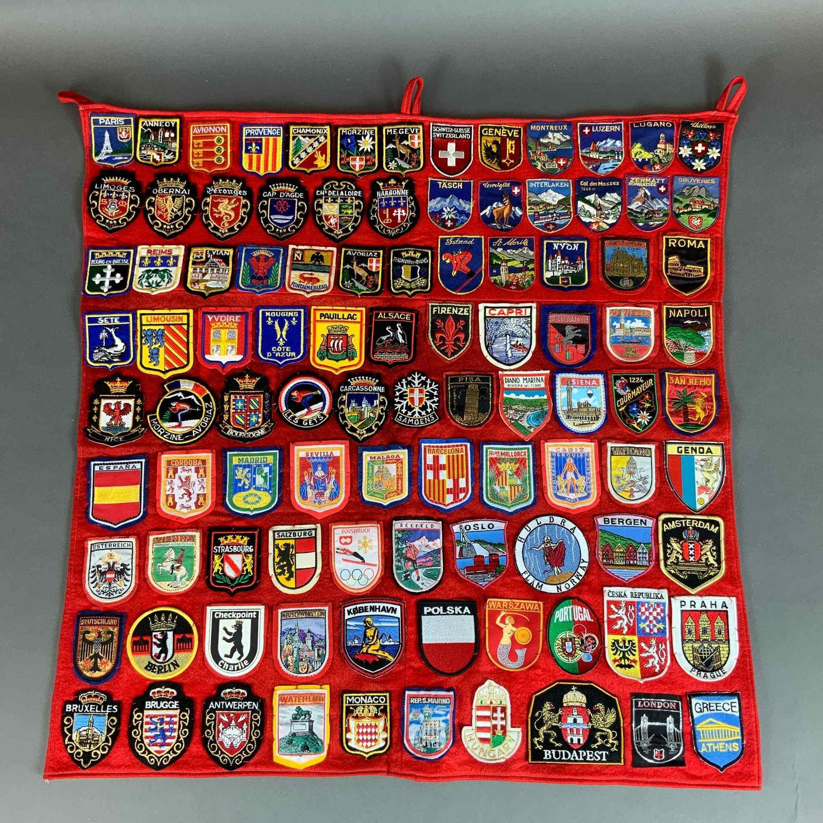 A Collection of Vintage European Travel Souvenir Patches Mounted on a Red Felt Banner: An assortment of approximately eighty vintage embroidered souvenir patches from various European countries, hand-sewn onto a rectangular red felt backing intended for wall display. The collection repr