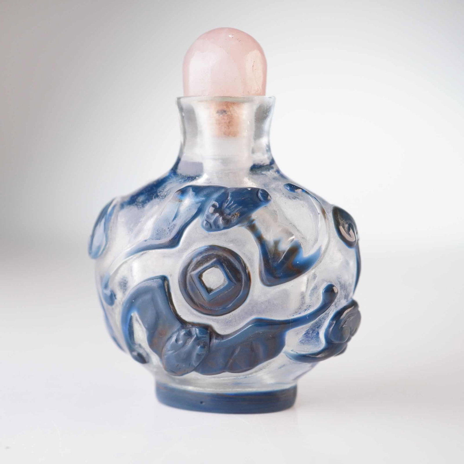 Peking Glass Snuff Bottle, Gray Overlay Bats, Coins, Ingots, and Lingzhi: Adorned with a gray relief overlay, this clear round Peking glass snuff bottle is topped with a rose quartz-hued stopper. It is decorated with depictions of bats, a coin, ingots, and lingzhi mushrooms