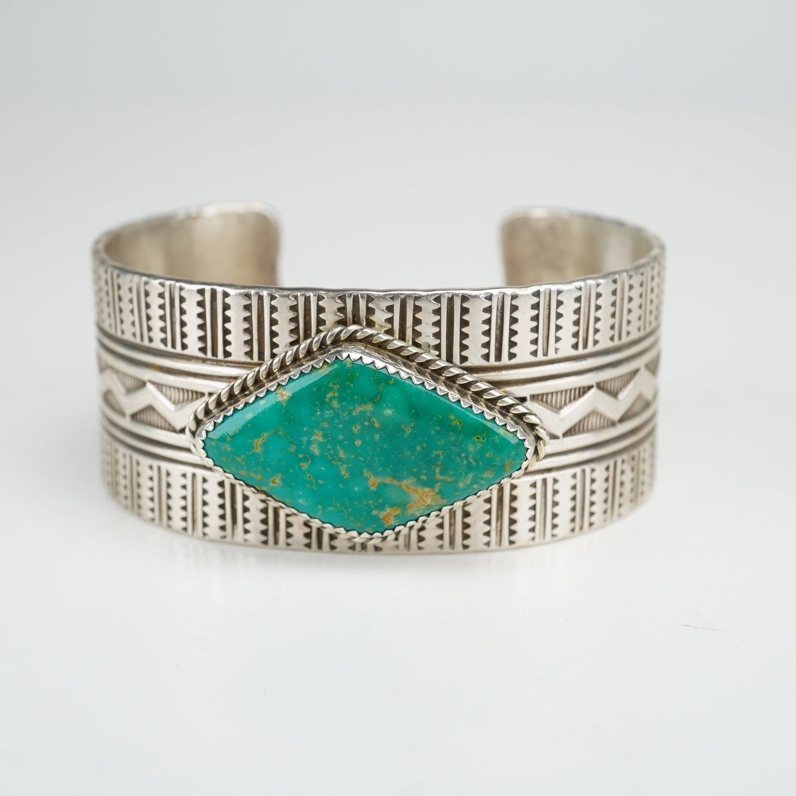 Navajo Sterling Silver and Turquoise Native American Cuff Bracelet Signed PJ: This Navajo handcrafted piece features a centrally set, diamond-shaped turquoise cabochon with a green-blue hue and a golden-brown matrix. The stone is secured within a sawtooth bezel and accented by 
