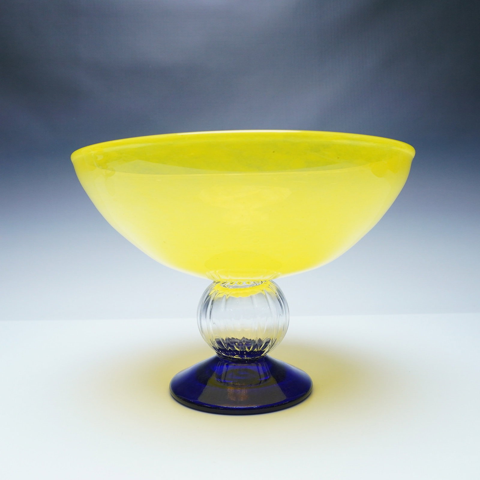 Kosta Boday by Gunnel Sahlin Centerpiece Bowl: A striking art glass bowl designed by Gunnel Sahlin for Kosta Boda. This sculptural piece features a vibrant yellow bowl contrasted with a cobalt blue foot, separated by a clear ribbed orb, creating a
