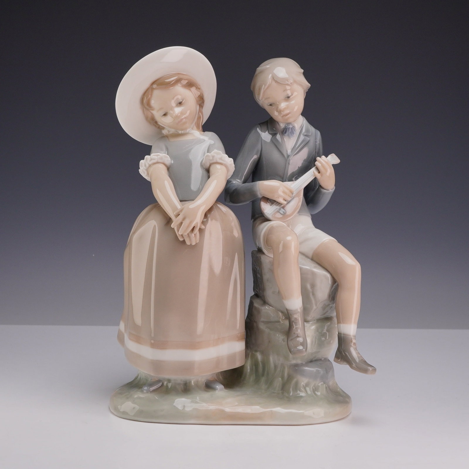 Lladro Porcelain Figurine, Adolescence 01004878: This Lladro glazed porcelain figurine, titled Adolescence and identified as model number 4878, depicts a pastoral scene featuring a young boy and girl. The boy is depicted seated upon a stacked stone 