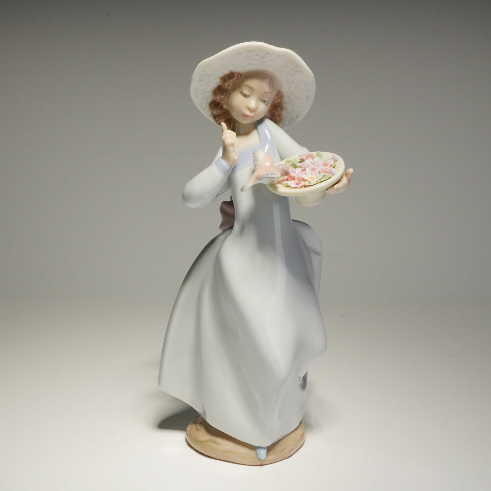 Lladro Porcelain Figurine Caught in the Act 1006439: A Lladro glazed porcelain figurine titled Caught in the Act, model number 1006439. The figure depicts a young girl wearing a wide-brimmed hat and a flowing white dress with a blue bodice and a large p