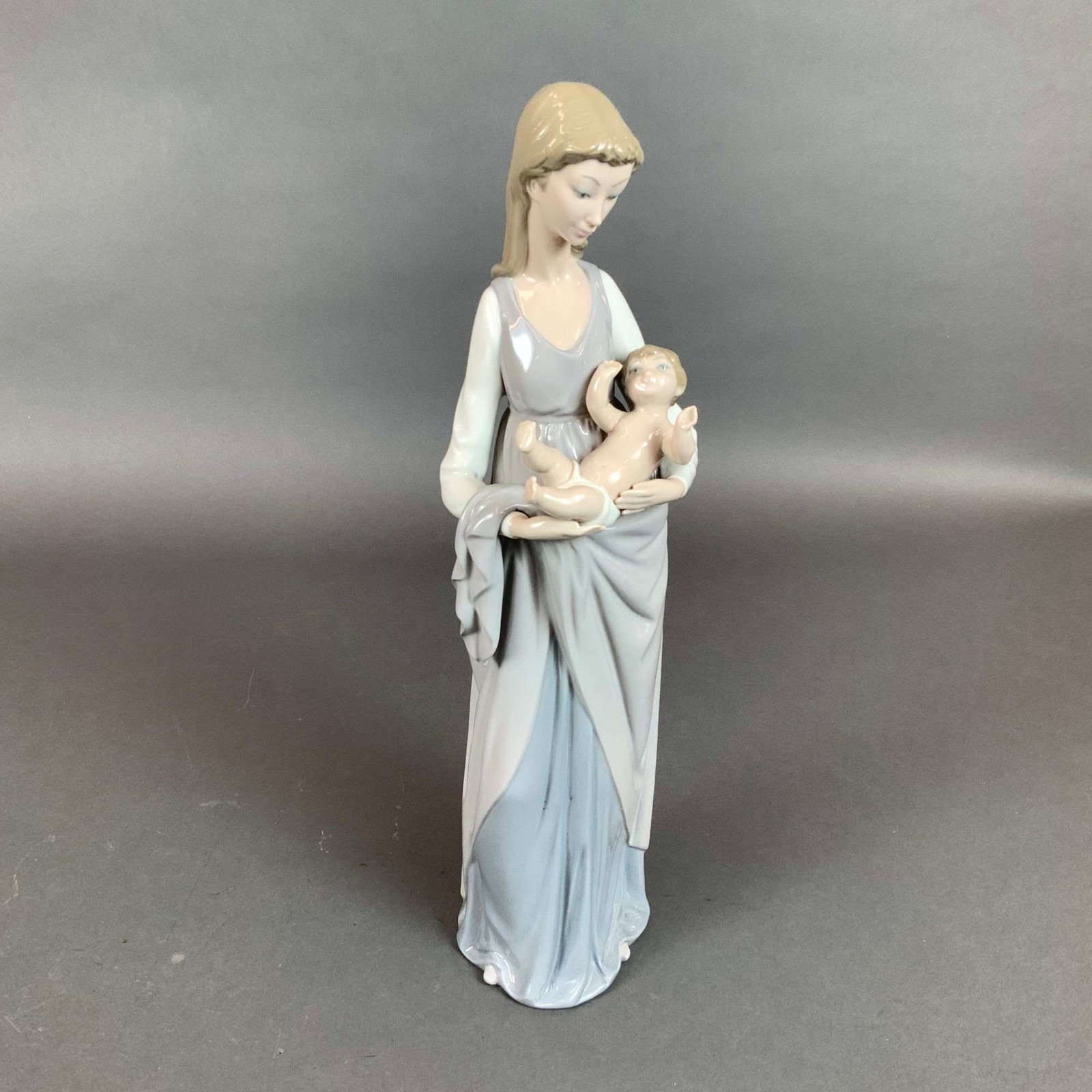 Lladro Porcelain Figurine Mother and Child Model Number 5086: This Lladro glazed porcelain figurine, model number 5086, titled Mother and Child. The piece portrays a standing woman in a long, flowing two-toned gown of pale gray and light blue, cradling a young c