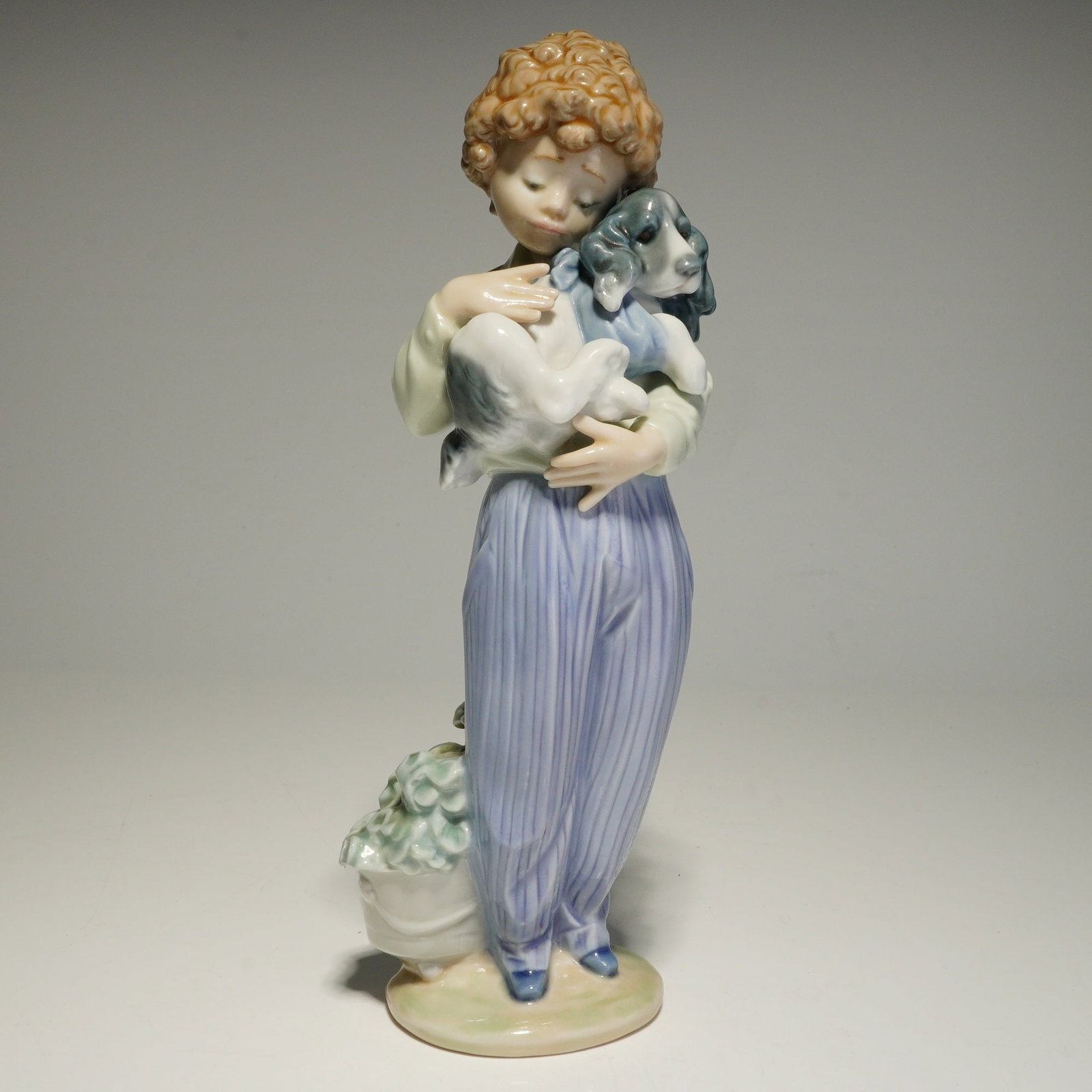 Lladro Porcelain Figurine, My Buddy 01007609: A Lladro glazed porcelain figurine issued for the 1989 Collectors Society. The sculpture depicts a young boy with curly brown hair holding a spaniel puppy. The figure is dressed in lavender-striped tr