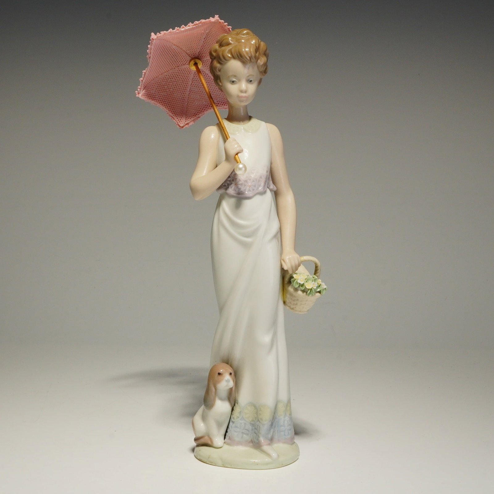 Lladro Porcelain Figurine, Garden Classic 01007617: The figurine depicts a young woman in a long white gown with floral accents at the waist and hem, holding a pink lace-patterned parasol and a basket of flowers, with a small dog seated at her side. Th