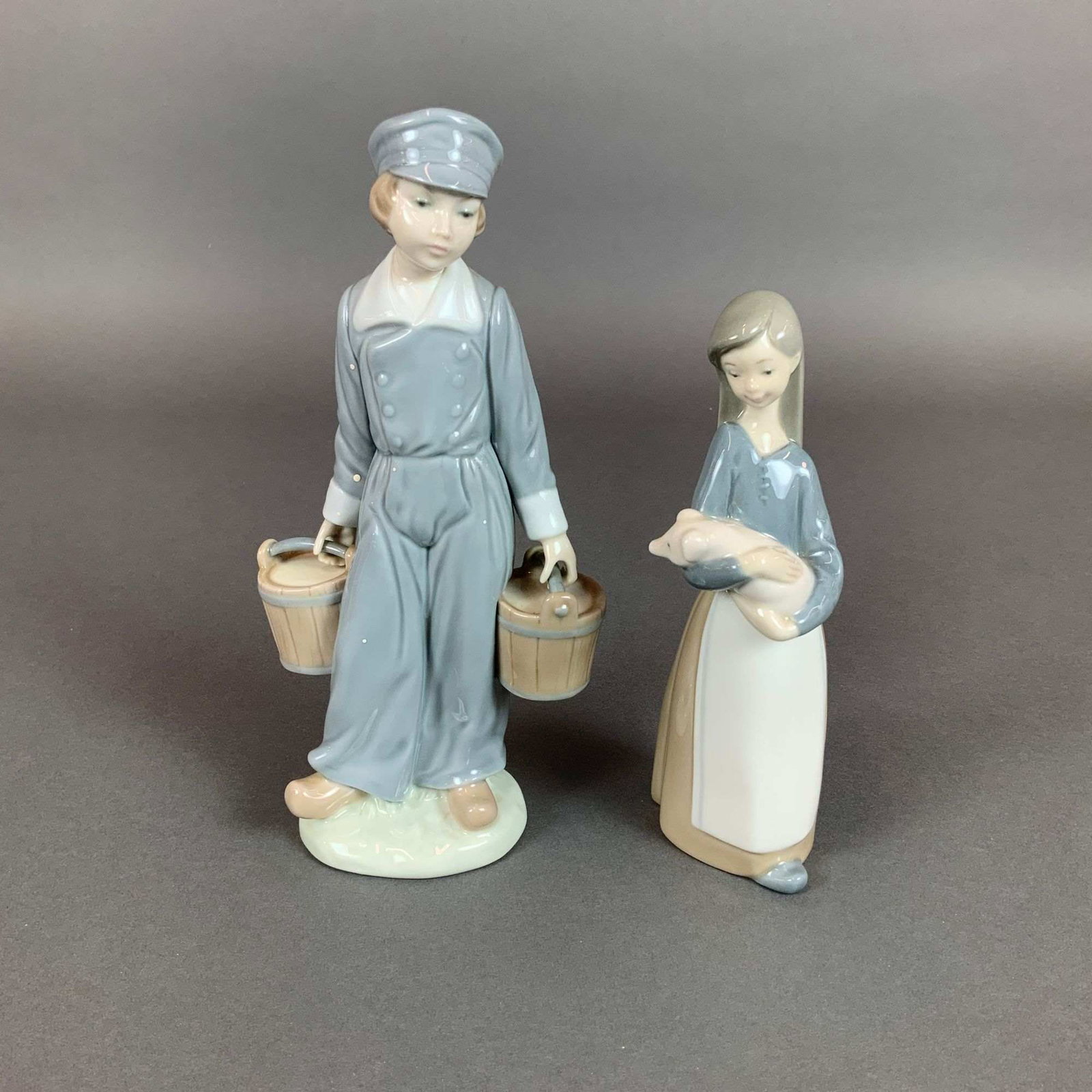 Two Lladro Glazed Porcelain Figurines, Boy with Pails and Girl with Pig: This lot features two glazed porcelain figurines manufactured by Lladro in Spain. The first figure, Boy with Pails model 4811, depicts a boy carrying two wooden-style buckets. He is dressed in a blue 