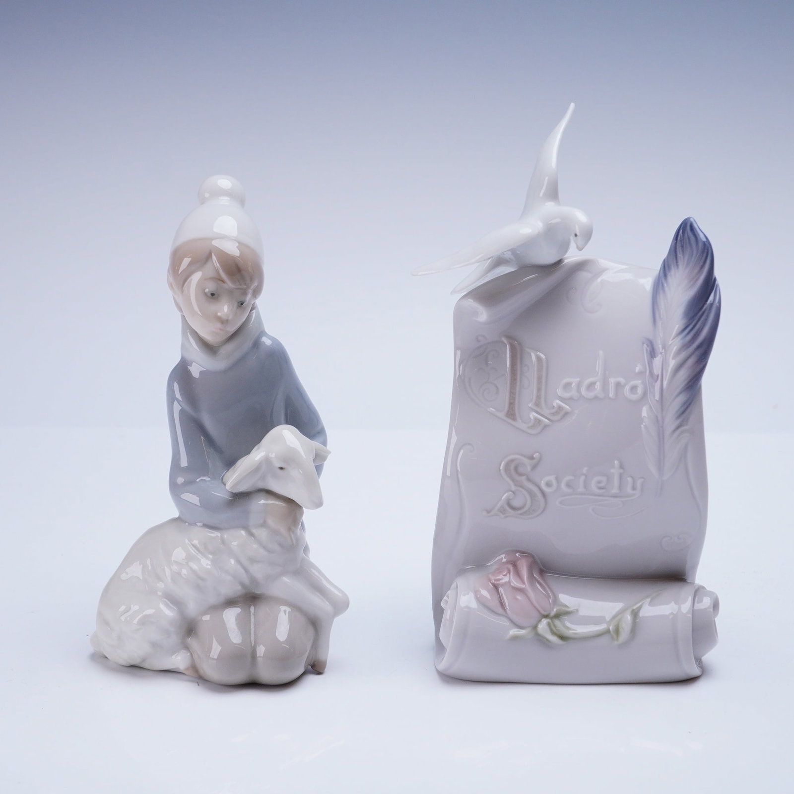 2pc Lladro Figurines Shepherd Boy with Lamb and Society Plaque: The lot includes the Lladro Shepherd Boy with Lamb figurine, identified by the model number 1004676), and a Lladro Society Plaque. The Shepherd Boy figurine depicts a young child kneeling and embracin