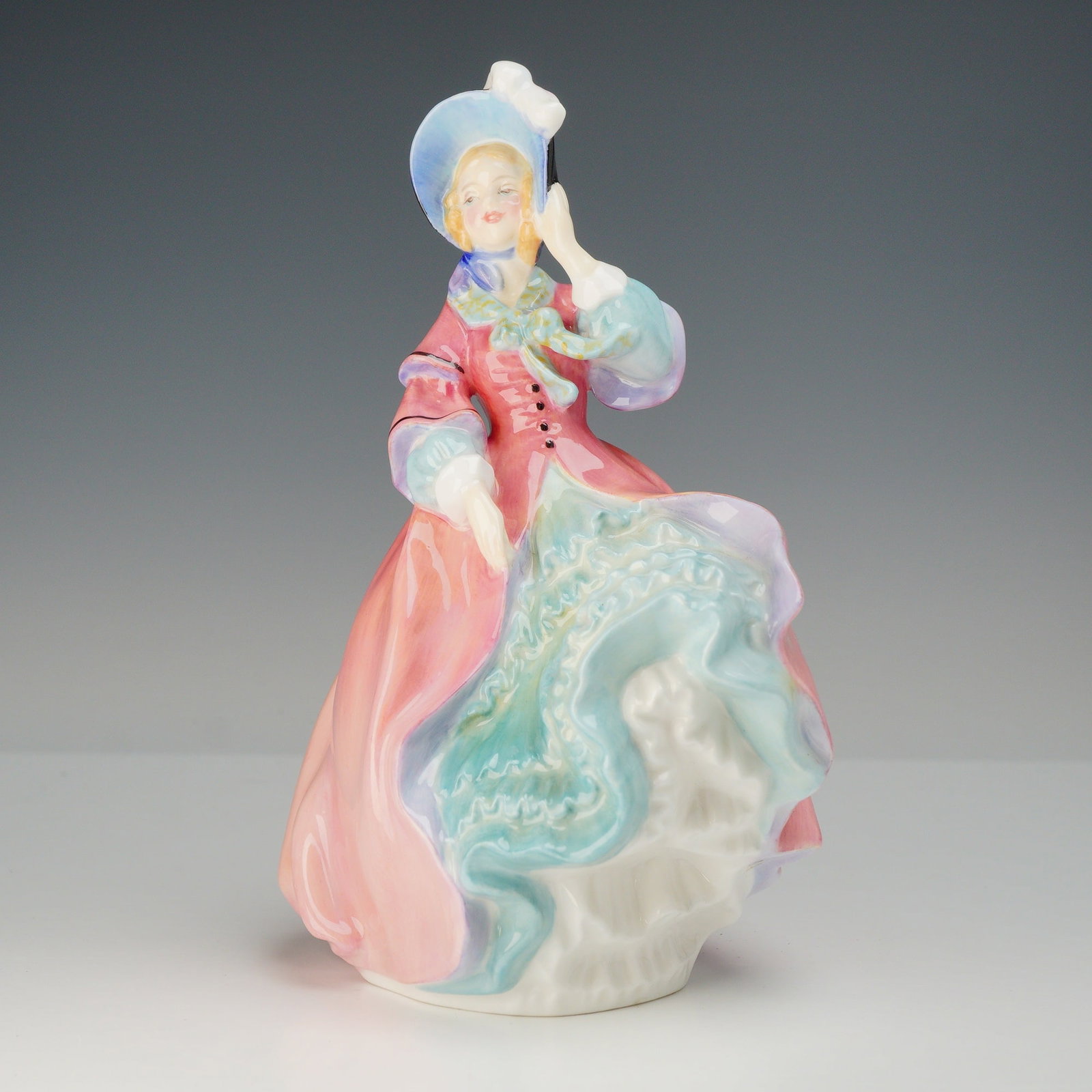Royal Doulton Bone China Figurine, Spring Morning HN1922: This hand-painted piece depicts a young woman in a windswept 19th-century style dress, featuring a billowing pink gown over a ruffled pale blue and white underskirt. She wears a blue bonnet and holds 