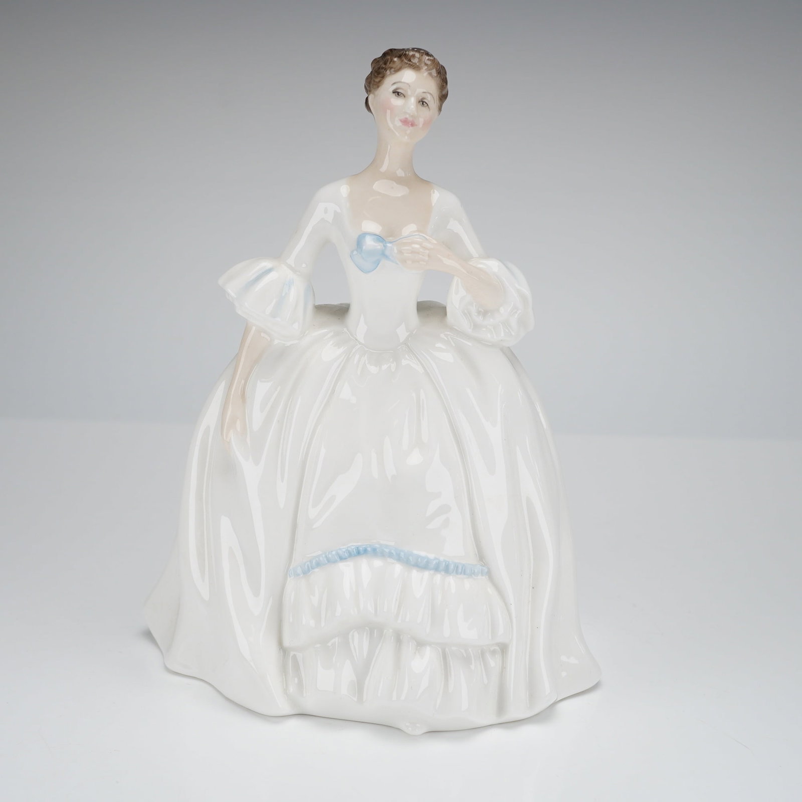Royal Doulton Kelly Porcelain Figurine HN3222: A single glazed porcelain figurine by Royal Doulton, identified as the "Kelly" model, HN 3222. The piece depicts a woman in a full-skirted, historical-style white gown, accented with light blue bows a