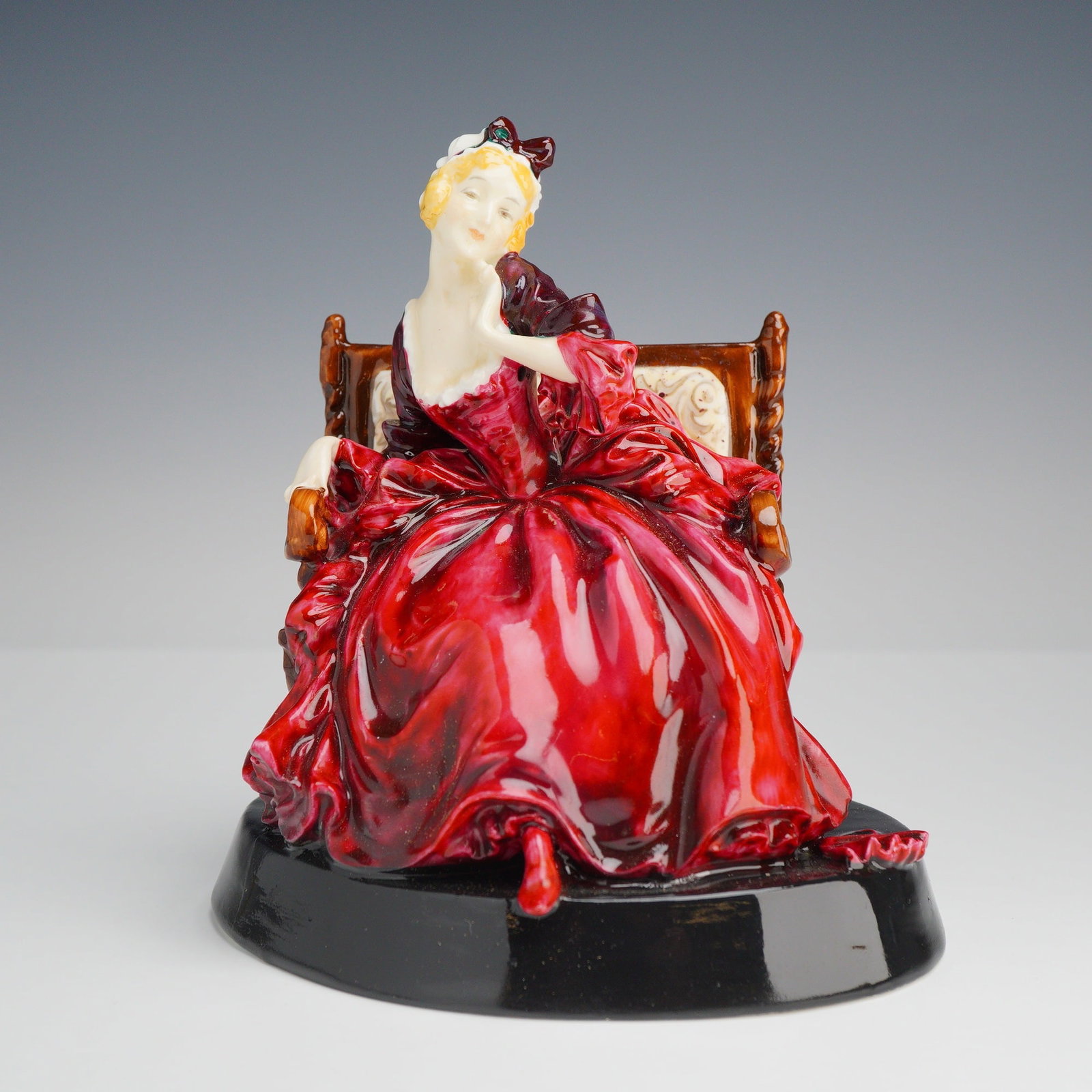 Royal Doulton Porcelain Figurine, Proposal Lady HN715: Depicts a woman with blonde hair wearing a white cap and dark bow. She is seated on a brown high-backed settee and wears a voluminous dress finished in a deep red glaze. The figure is posed with a til
