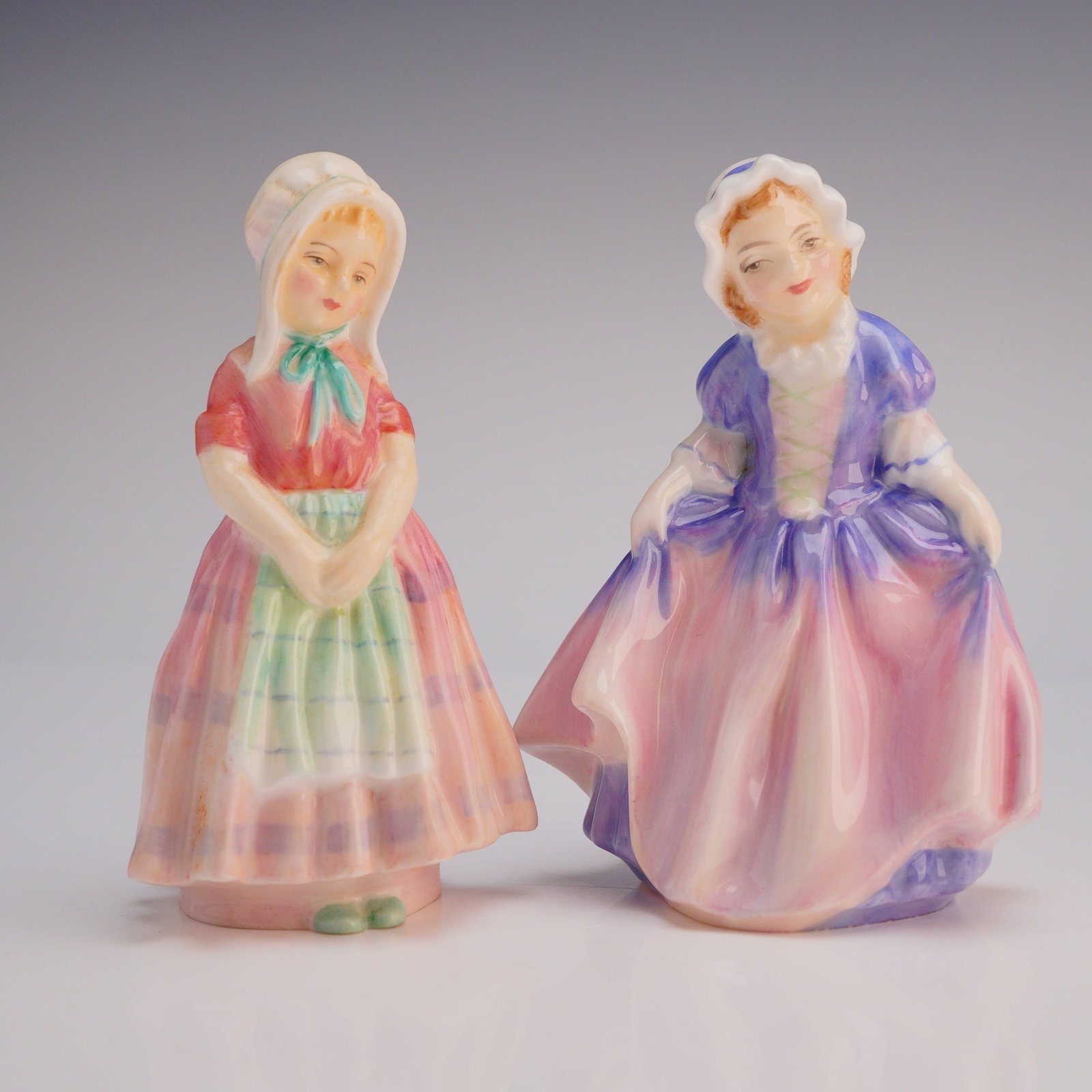 2pc Royal Doulton Figurines Dinky Do HN1678 and Tootles HN1680: The figures are executed in high-gloss glazed porcelain with polychrome enamel decoration, utilizing pastel tones of pink, purple, red, and green. They depict young women wearing bonnets and period co
