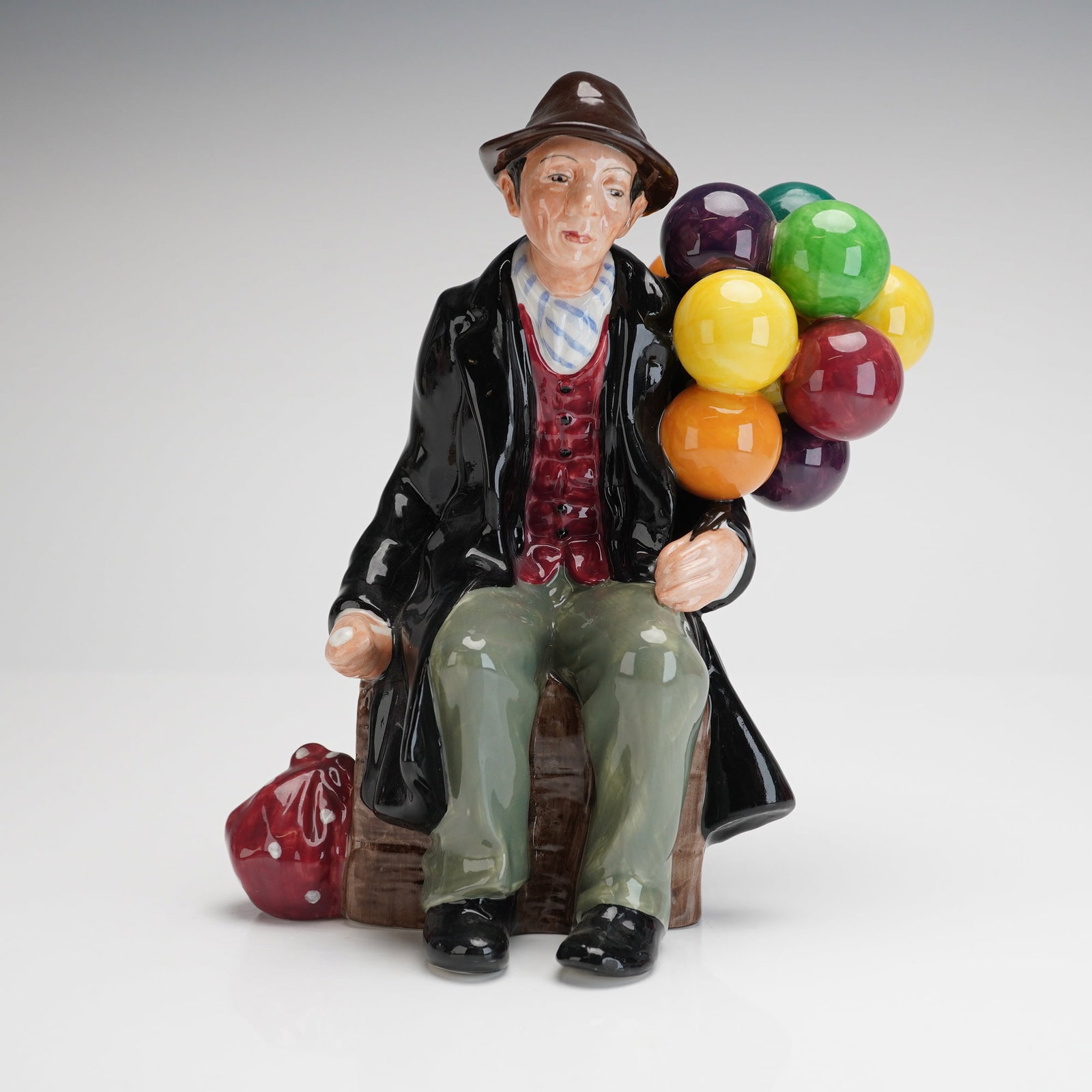 Royal Doulton Figurine The Balloon Man HN1954: The polychrome figure depicts a seated street vendor, wearing a brown fedora and a black overcoat over a red waistcoat and green trousers. The vendor holds a large cluster of multicolored balloons, in