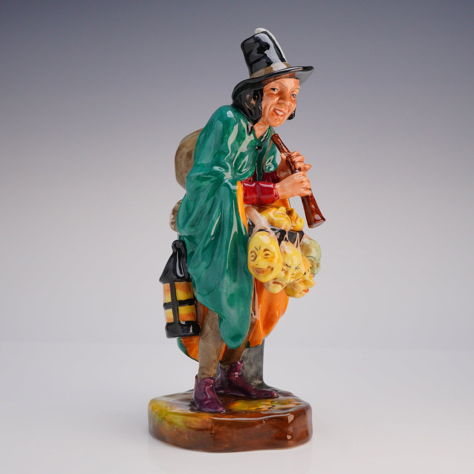 Royal Doulton Porcelain Figurine The Mask Seller HN2103: The figure is attributed to designer Leslie Harradine. The polychrome enameled figure depicts a male street vendor or entertainer, modeled standing while playing a woodwind instrument. He is dressed i