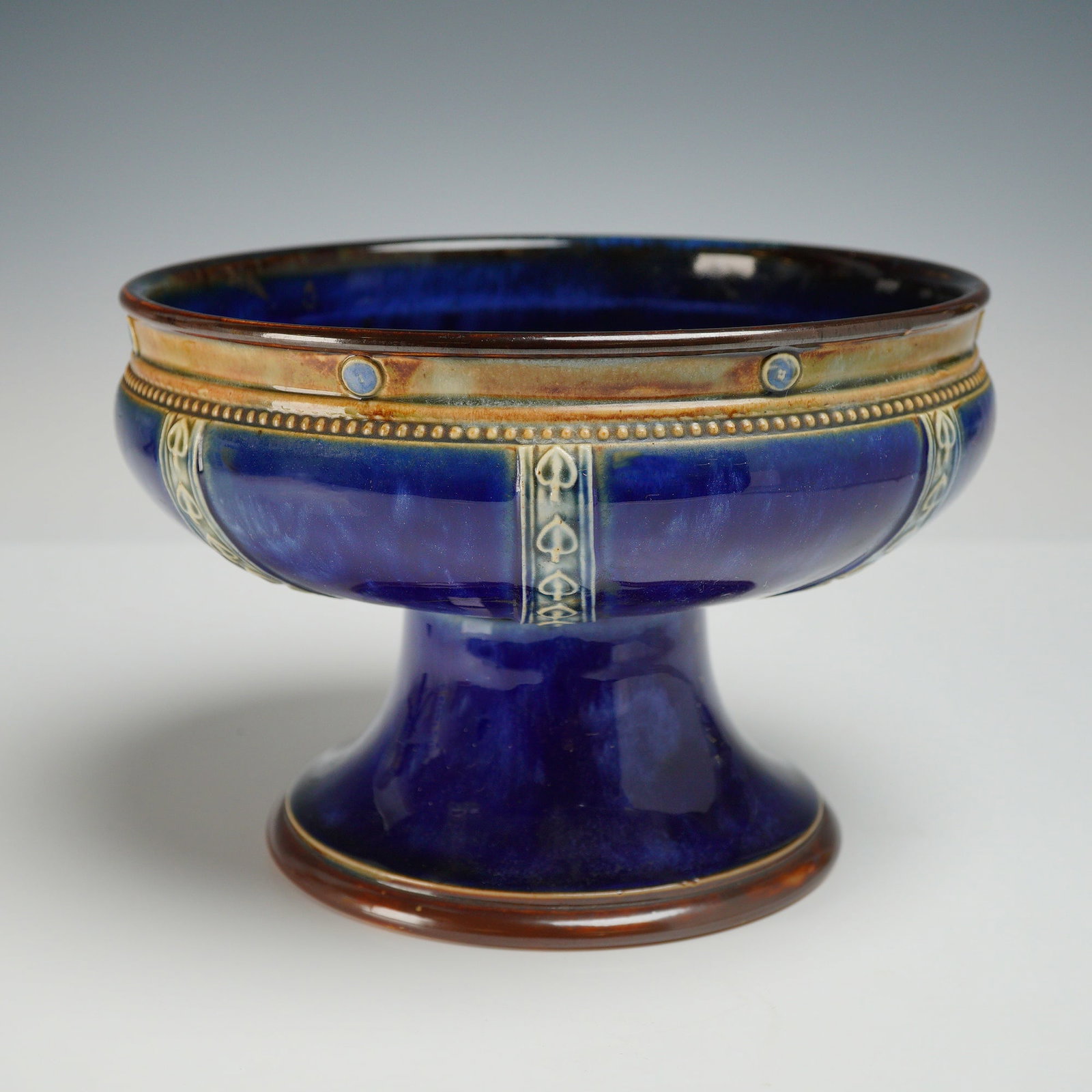 Royal Doulton Cobalt Blue Glazed Stoneware Footed Bowl: This Royal Doulton glazed stoneware footed bowl features a wide, shallow basin supported by a flared pedestal base. The exterior is finished in a deep, mottled cobalt blue glaze, while the rim is acce