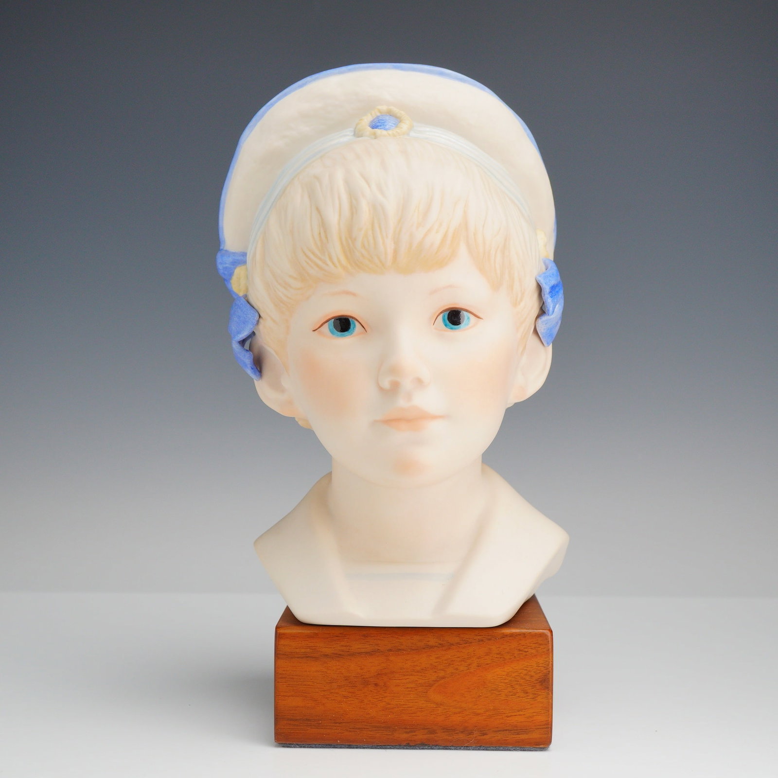Cybis by Gertrude Fass Porcelain Bust, Edward: This porcelain bust depicts a young Victorian-era boy named Edward, shown with short blond hair and blue eyes. He wears a white sailor-style hat with blue trim and matching blue side ribbons. The ungl