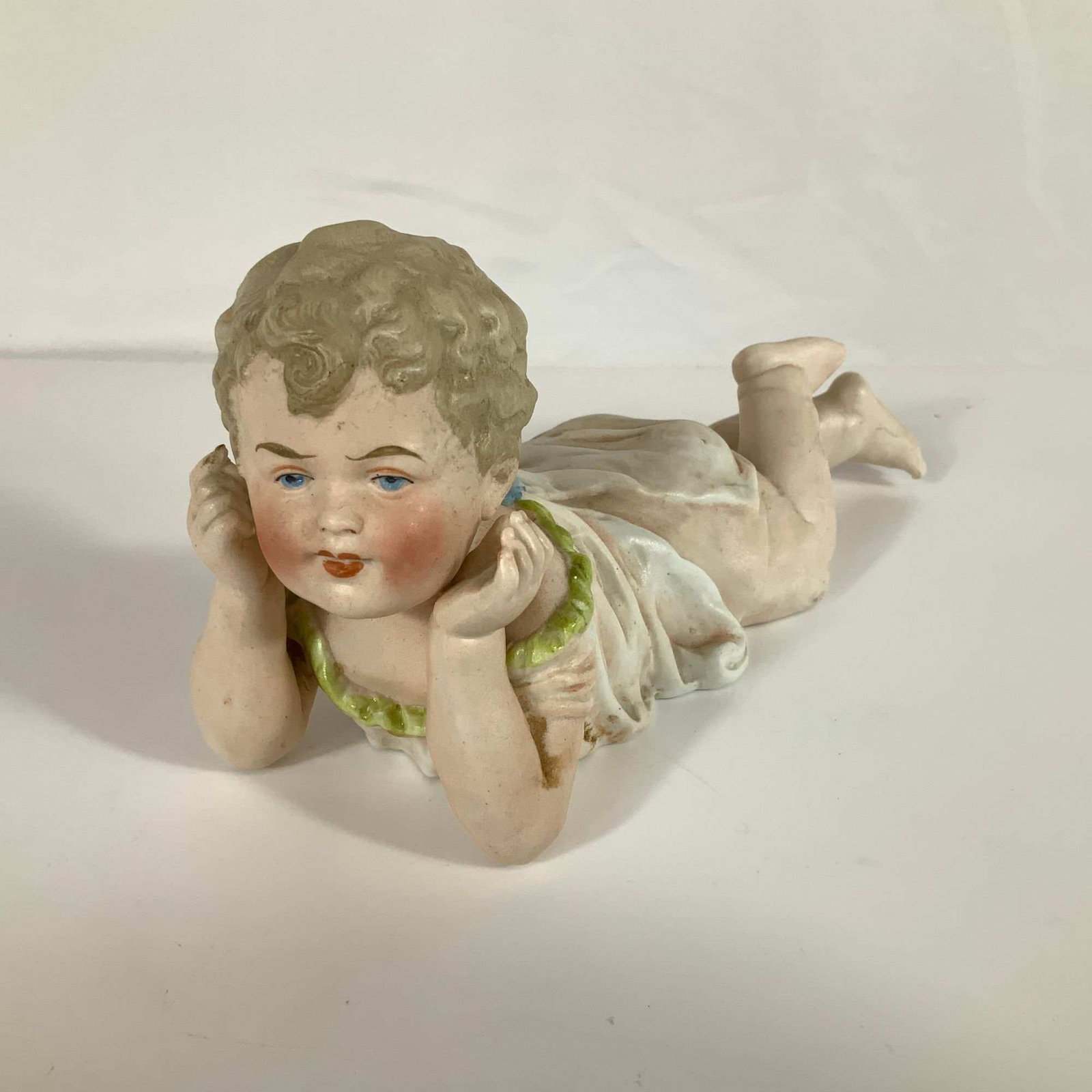 German Bisque Porcelain Piano Baby Figurine Model 484: A hand-painted bisque porcelain Piano Baby figurine depicting a young child in a prone position. The infant is shown lying on its stomach with its head resting near its hands. The figure wears a white