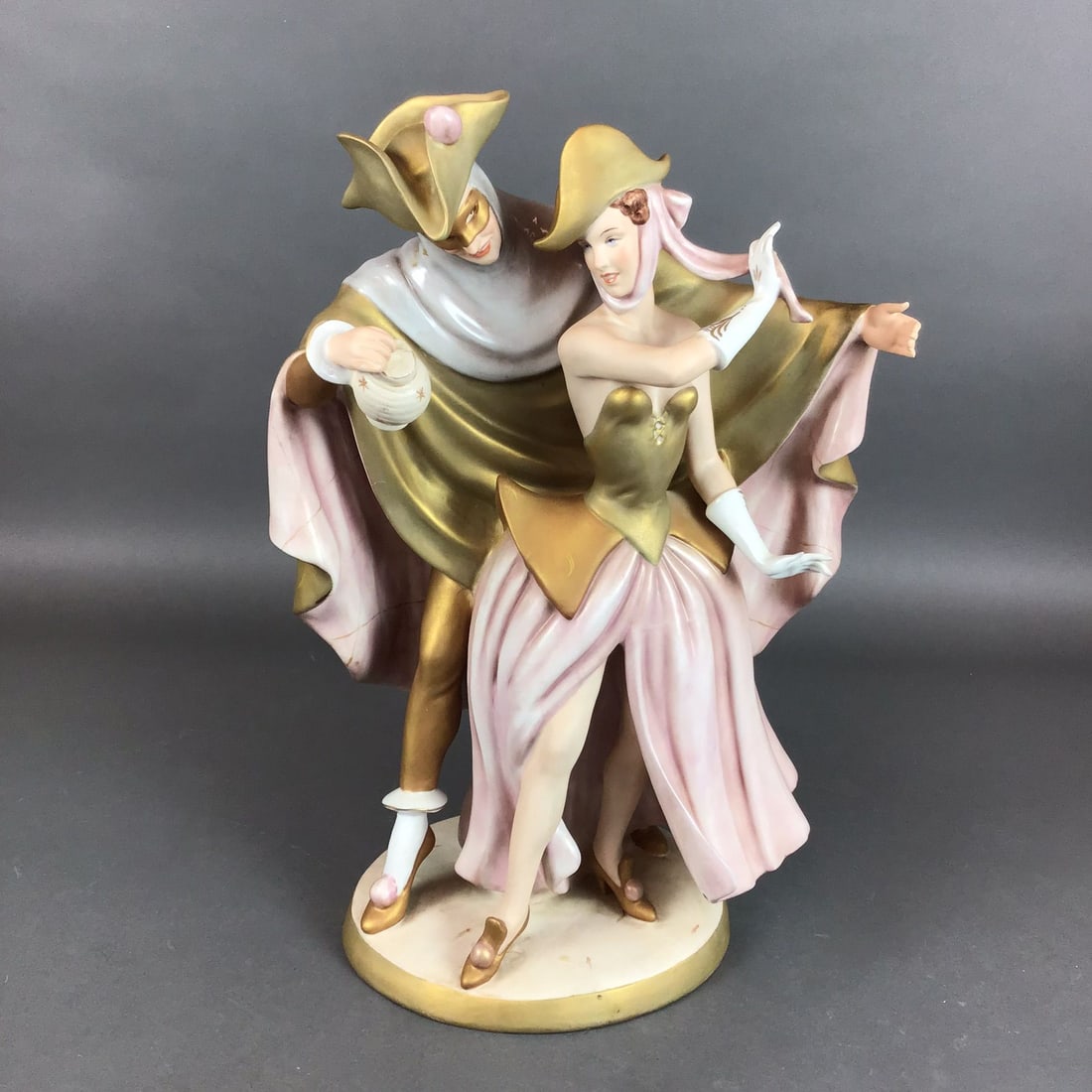 Royal Dux Art Deco Porcelain Masquerade Dancing Couple Figurine: This Royal Dux porcelain figurine depicts a couple in stylized masquerade attire. The male figure is featured in a golden tricorne hat with a pink pom-pom, a mask, and a voluminous draped cape in gold