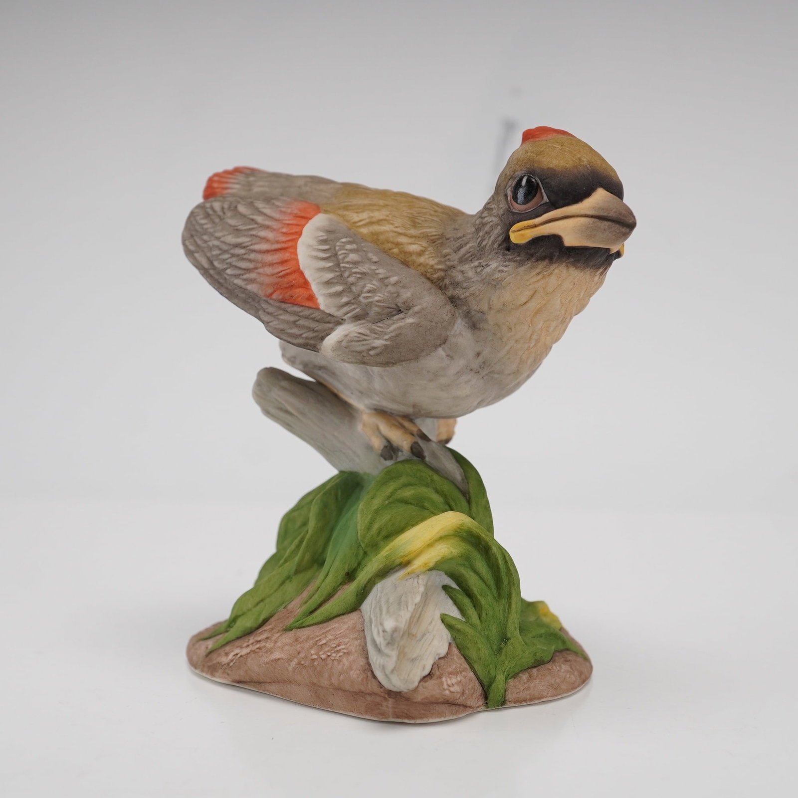 Boehm Porcelain Figurine, Baby Cardinal 400-57: This hand-painted matte bisque porcelain piece depicts a fledgling bird in a lifelike pose, perched upon a textured branch that rises from a cluster of green foliage. The cardinal features brownish-gr