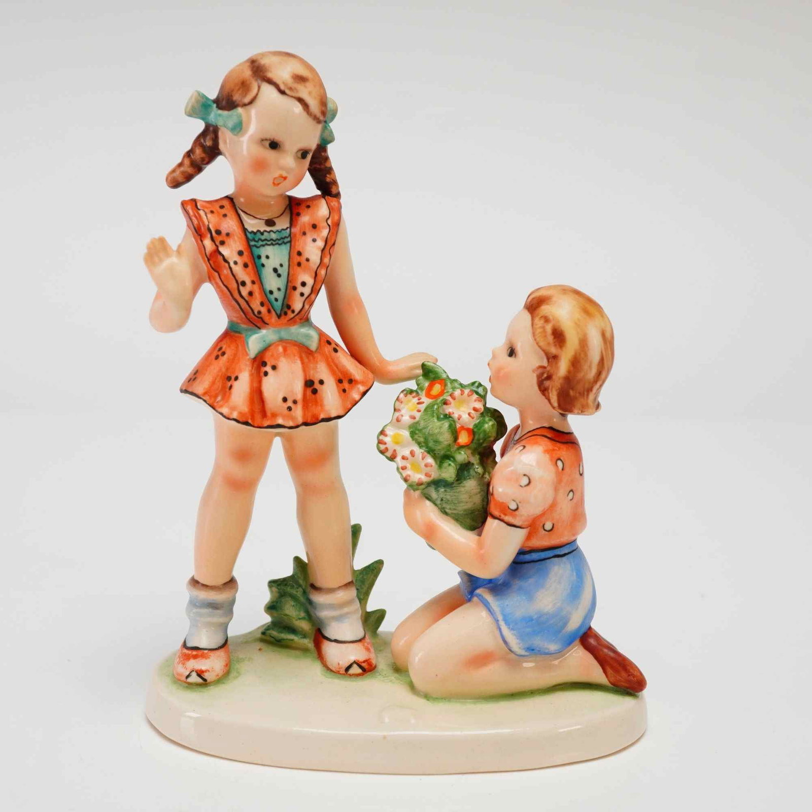 Goebel Nasha We Congratulate Hum220 Porcelain Figurine: A Goebel Nasha glazed porcelain figurine titled We Congratulate, model Hum 220. The piece depicts two young children in mid-century attire, featuring a standing girl with pigtails in a spotted orange 