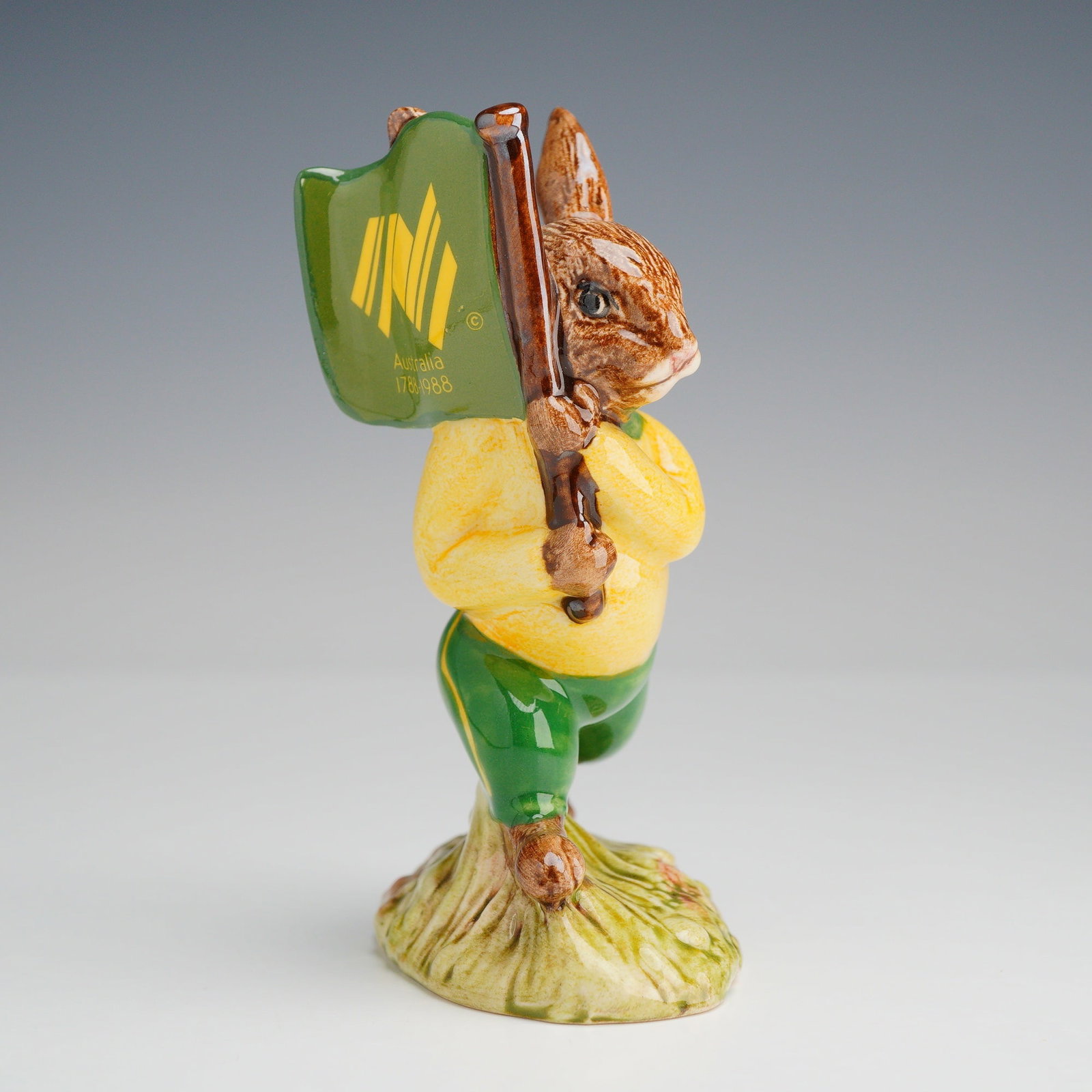Royal Doulton Bunnykins Figurine, Australian Bunnykins DB58: The glazed ceramic anthropomorphic rabbit is depicted in a walking pose, wearing a yellow shirt and green trousers with yellow side stripes. The figure holds a green flag featuring the yellow graphic 