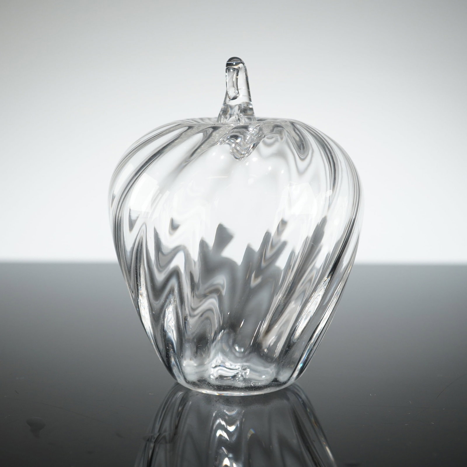 Crystal Holiday Apple Ornament / Paperweight: The object is formed from clear, colorless glass, functioning as a paperweight or small sculpture. The body features a pronounced twist or swirl-optic pattern that wraps dynamically around the apple f