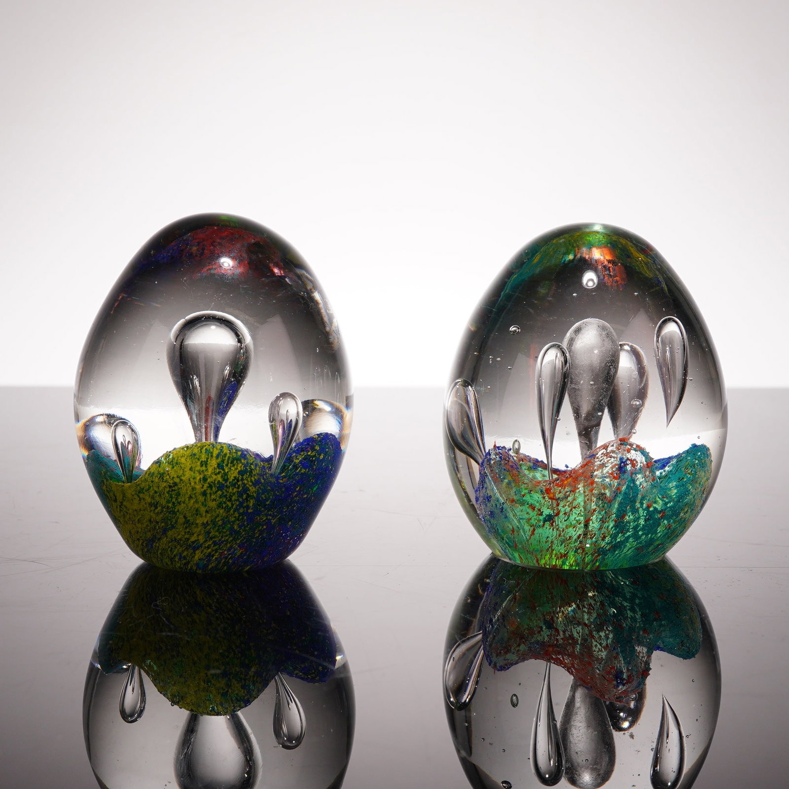 2pc Art Glass Controlled Bubble Egg-Shaped Paperweights: Each piece is hand-worked from heavy, solid clear glass and features internal multicolored frit decoration at the base. One weight displays a palette of blue, yellow, and green crushed glass, while th