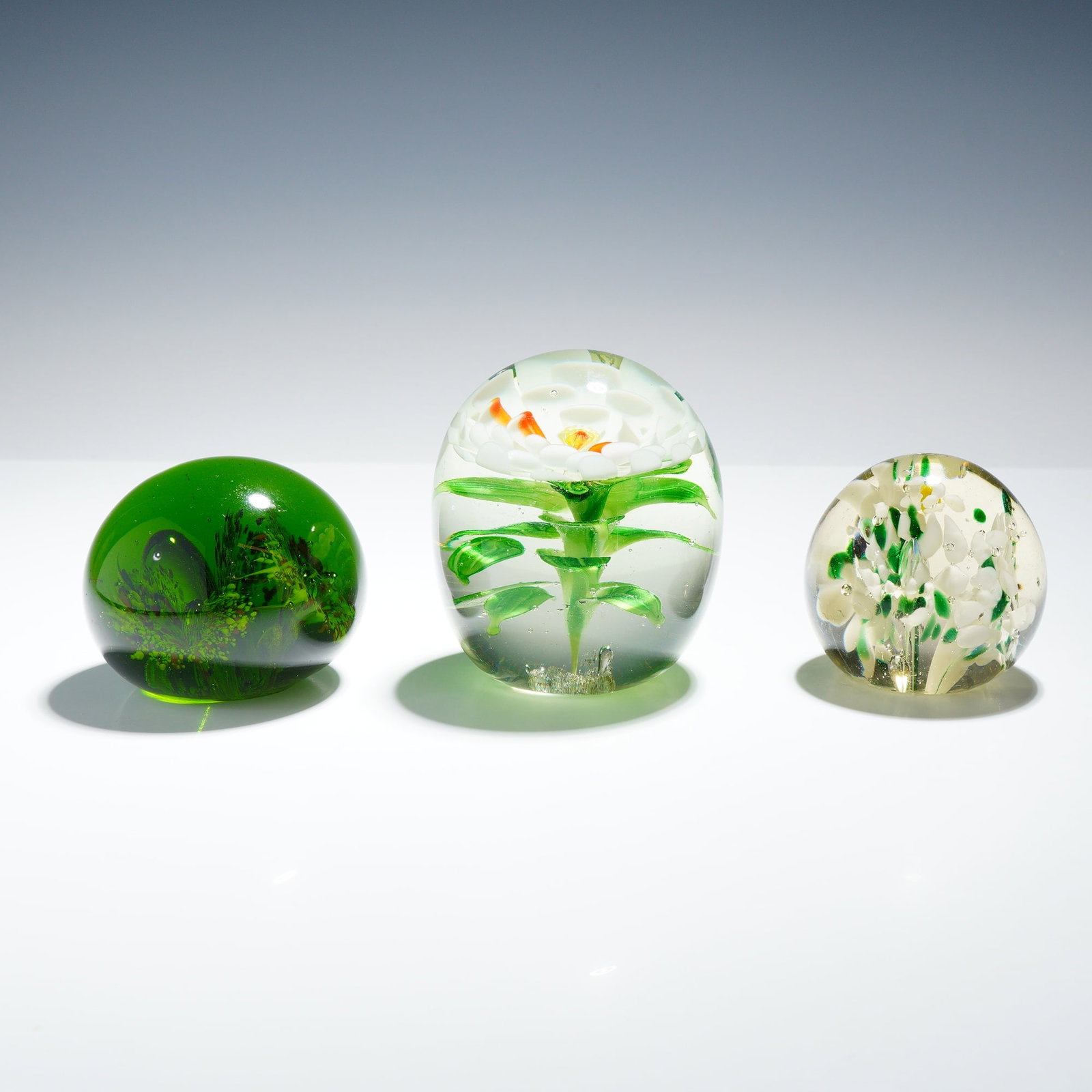 3pc Art Glass Paperweights with Floral Themes: This 3pc collection includes three decorative art glass paperweights featuring various internal lampwork and controlled-bubble techniques. The largest item is an ovoid clear glass paperweight containi