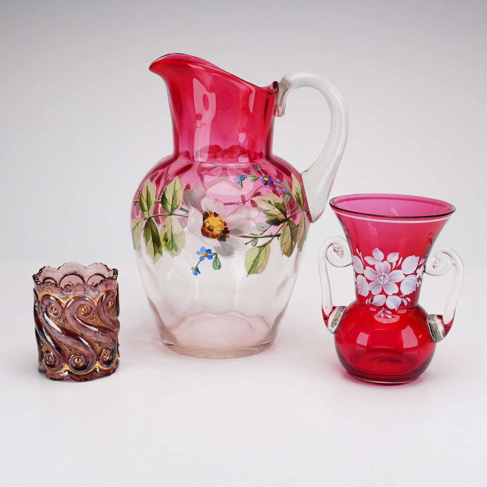 3pc Decorative Art Glass Grouping: This three-piece collection of decorative art glass features varied techniques and hand-painted enameled decorations. The first item is a hand-blown Rubina glass water pitcher, transitioning from a cr