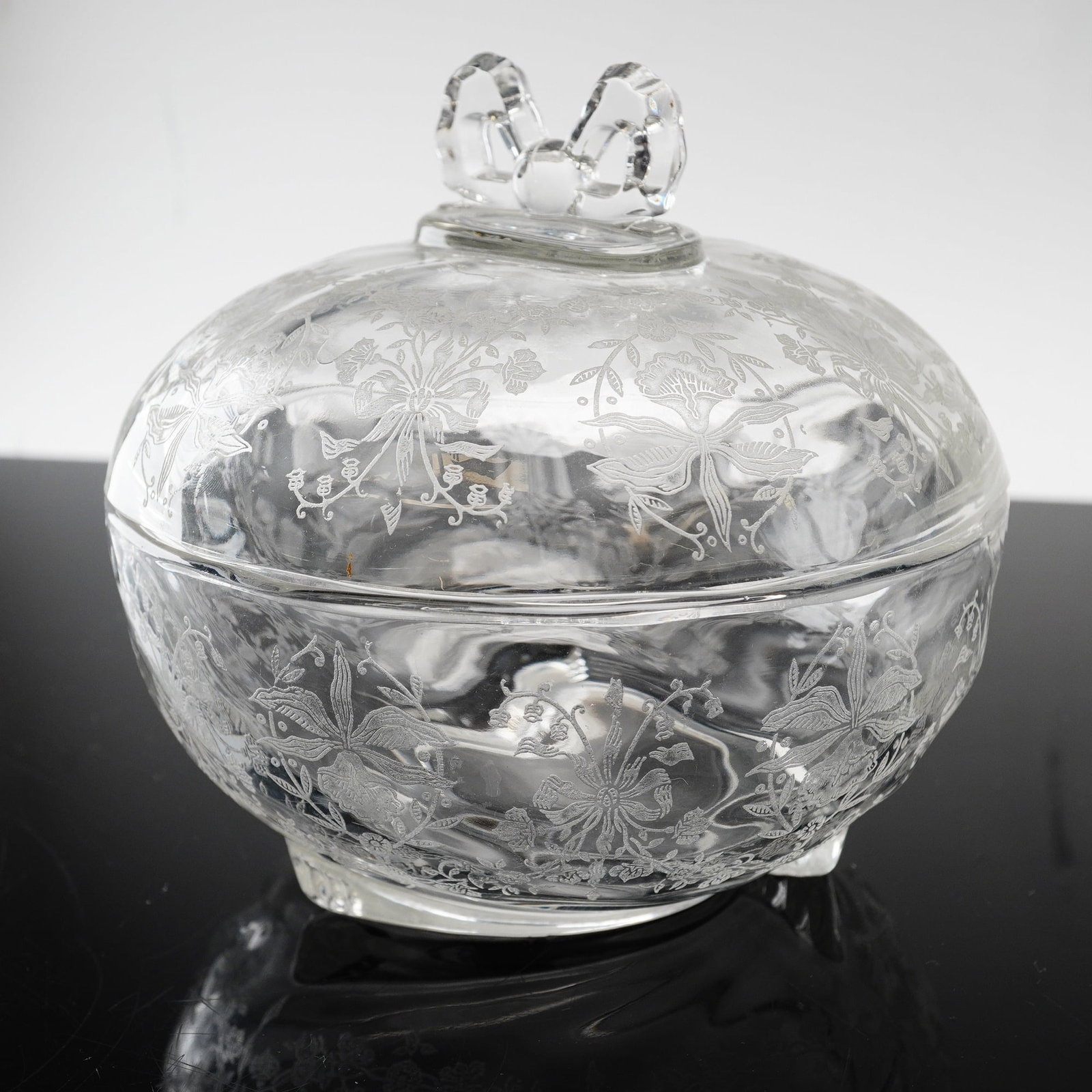 Heisey Orchid Etched Glass Covered Candy Dish with Bow Finial: This two-piece covered candy dish is manufactured by A.H. Heisey and Company of Newark, Ohio, featuring the Orchid etching. The piece is constructed of clear pressed glass with an acid-etched floral m