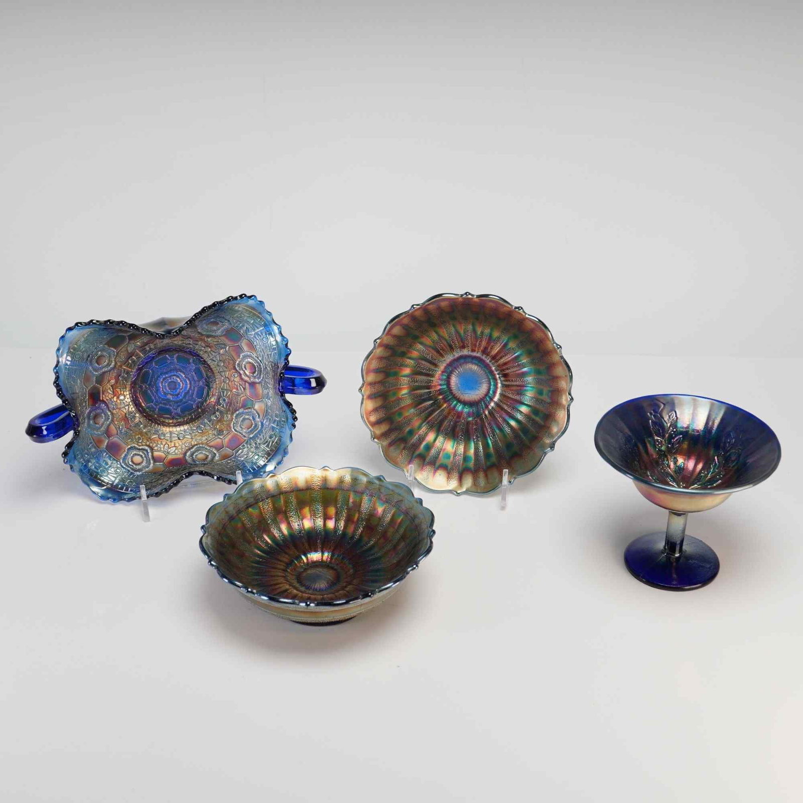 Four Cobalt Blue Carnival Glass Tableware Articles: This group comprises four pieces of pressed carnival glass featuring a cobalt blue base with iridescent metallic salt finishes. The collection includes a two-handled bonbon dish attributed to Northwoo
